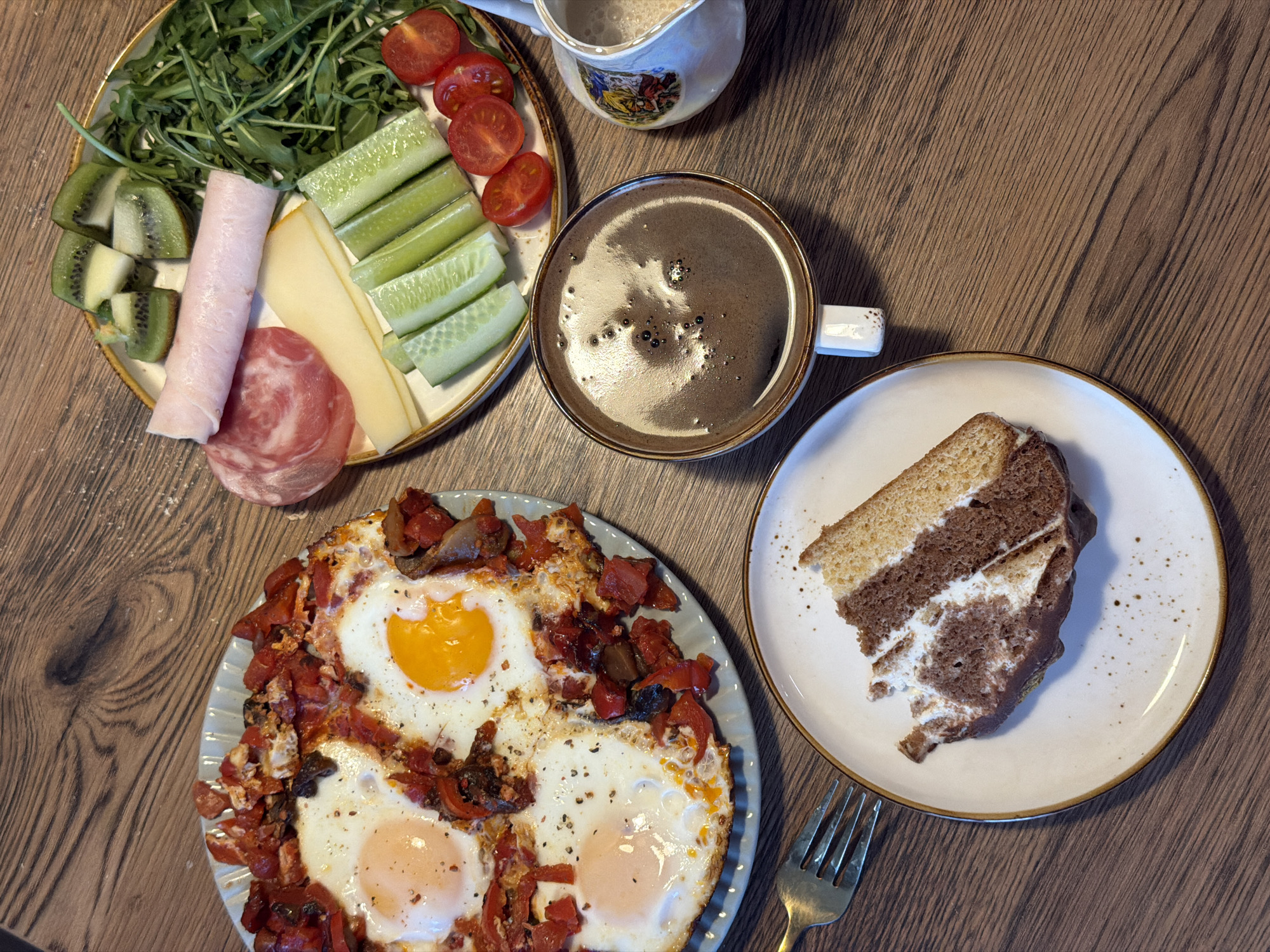An overhead view captures a varied meal laid out on a dark brown wooden table, in a location in Perm, Russia. The scene features multiple dishes for what appears to be a breakfast or brunch.
On the left, a light blue plate holds a dish resembling shakshuka, with four fried eggs nestled among diced red bell peppers and other dark vegetables. Each egg has a distinct yolk, with one yolk being a brighter orange. Black pepper flakes are visible on the eggs.
To the right, a white plate with a gold rim displays an assortment of cold items: several slices of green kiwi fruit, a mound of fresh arugula, two rolled slices of pink ham, a round slice of salami, two rectangular slices of light yellow cheese, multiple vertically cut cucumber sticks, and several halves of red cherry tomatoes.
Below these, a white ceramic mug with a brown rim contains a dark beverage, likely coffee, topped with a layer of foam and small bubbles. Adjacent to the mug, a small white creamer or milk pitcher with colorful, abstract designs on its side is visible, containing a light-colored liquid.
In the bottom left, a white plate with a gold rim holds a partially consumed slice of marble cake. The cake features distinct layers of light yellow sponge and dark brown chocolate sponge, interspersed with white cream filling. The base of the cake slice is covered in a dark chocolate frosting. A gold-colored fork rests on the table, to the left of this plate.
The wooden table surface shows prominent grain patterns throughout the image. No individuals are present in the frame.