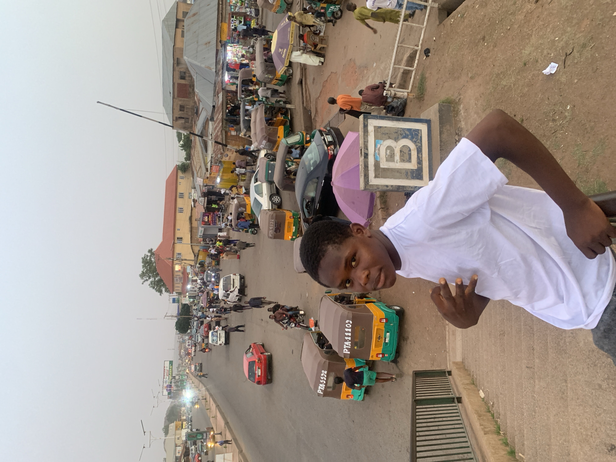 This image depicts a bustling street scene in an urban area, consistent with daily life in Jos, Nigeria, viewed from an elevated position. In the foreground, on the right, a young man with dark skin, wearing a white t-shirt and grey trousers, poses for the camera with a slight smile. He appears to be standing on a pedestrian bridge or a set of stairs.
Below and extending into the mid-ground is a busy street. The road is populated by numerous vehicles, including several green and brown auto-rickshaws (known as Keke Napeps), with registration numbers like "PTA 5572," "PTA 11103," and "PTA 41103" visible on their rear. Cars, such as a red sedan, a black car, and a white minivan, are also part of the traffic flow. People are seen walking along the roadside and congregating around market stalls.
Along the street, various buildings are visible, some with corrugated iron roofs and others, like a prominent building with a reddish-brown roof, suggesting residential or commercial structures. Market activity is evident, with vendors and customers amidst stalls, some shaded by umbrellas, including a distinct purple one and a yellow one with the partially visible text "MUMU." Near the young man, a large blue and white sign prominently displays the letter "B." A black and white barrier pole extends across part of the street in the middle distance.
The sky appears overcast or hazy, suggesting either a cloudy day or possibly late afternoon lighting. The overall scene captures a lively, active urban environment characterized by commerce, pedestrian movement, and vehicle traffic.