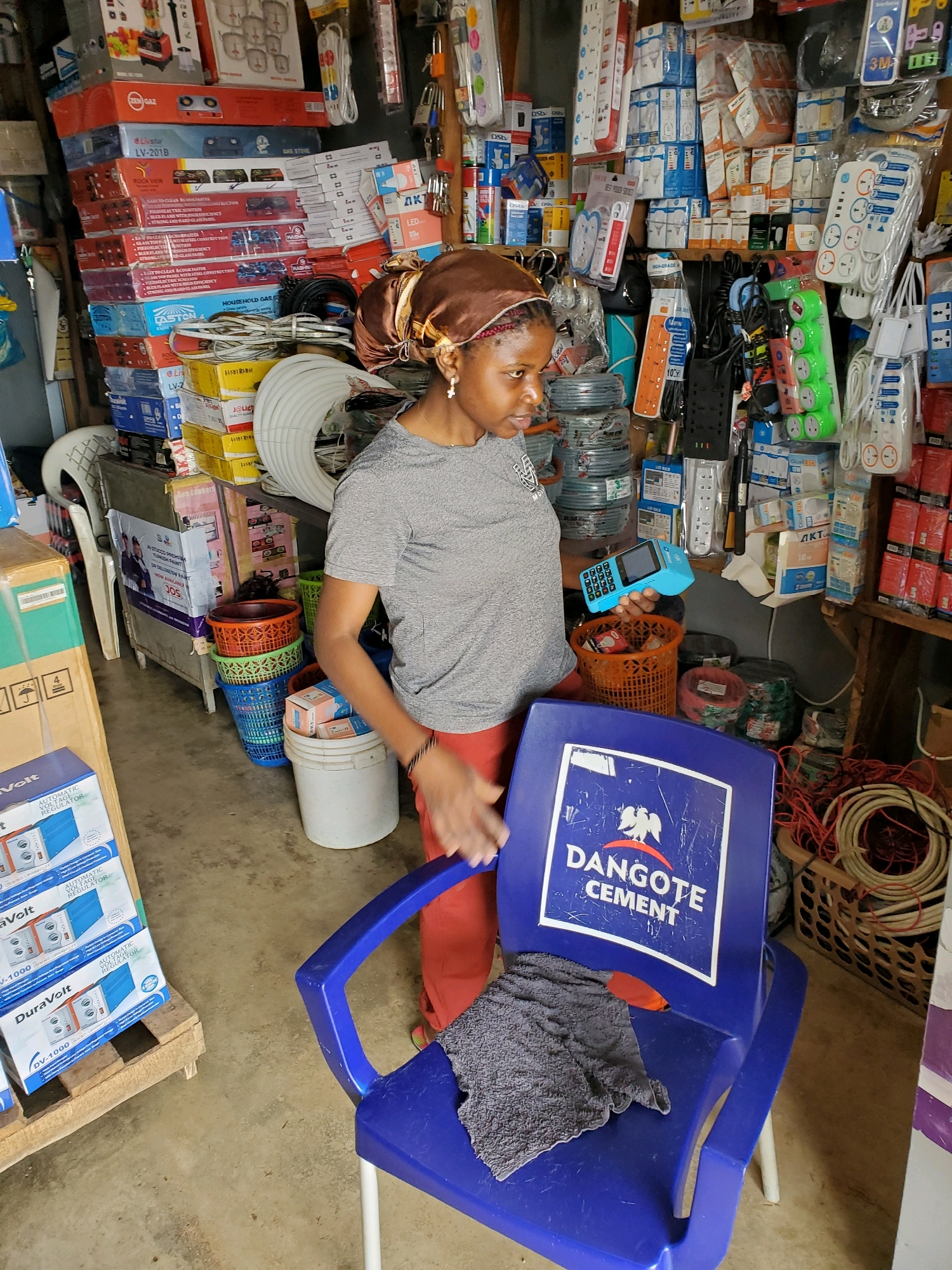 The image shows a woman standing in a retail store in Gura Top, Nigeria, holding a blue point-of-sale device. The store is filled with various electrical supplies, including extension cords, power strips, and boxes of appliances. Stacks of products are visible on shelves and on the floor. A blue plastic chair with the "Dangote Cement" logo sits in the foreground. The woman is wearing a grey t-shirt, orange pants, and a headscarf. The scene suggests a typical commercial environment where transactions are conducted.