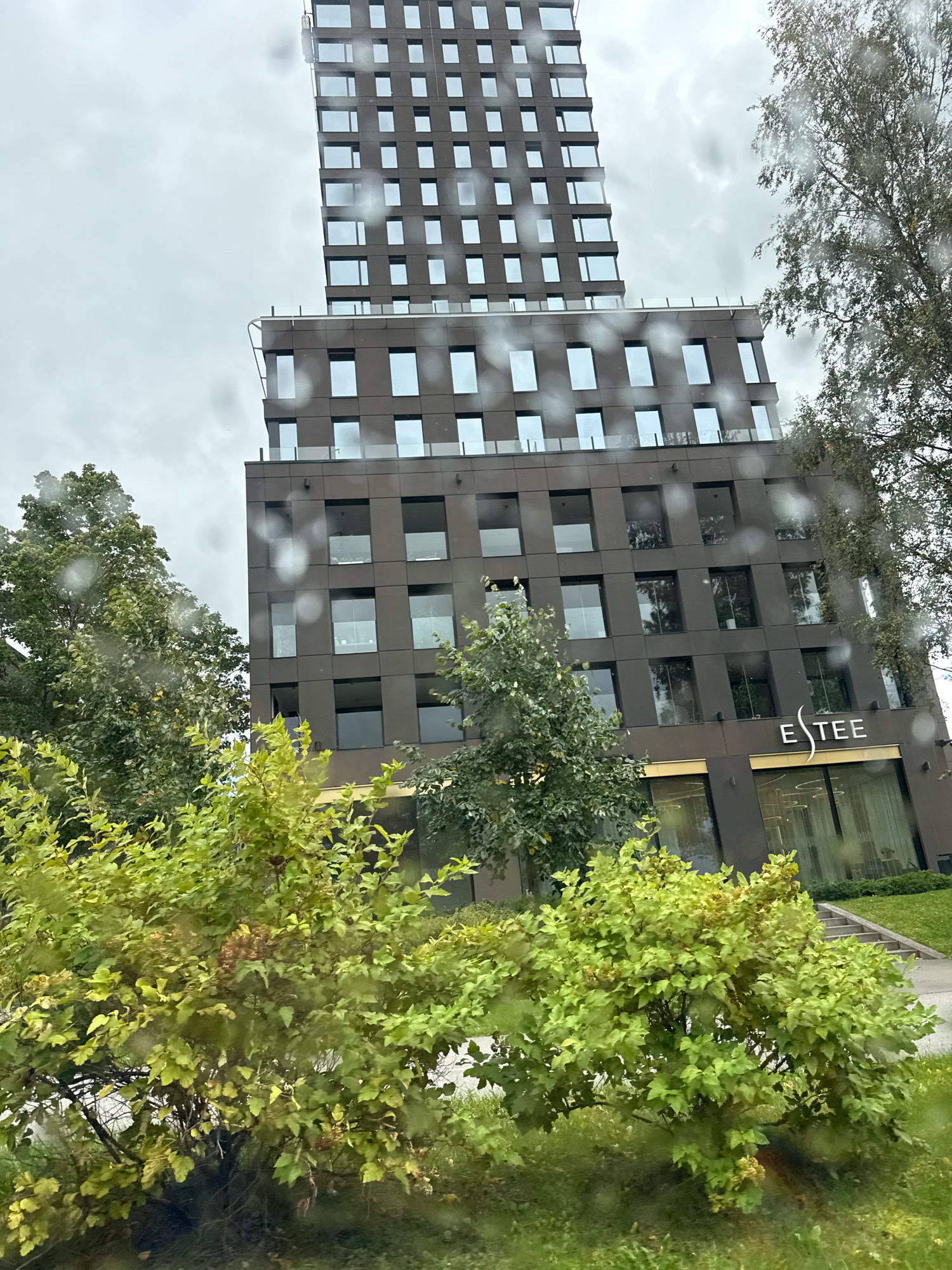 The image shows a modern, multi-story building with a dark facade and numerous windows, located in the city of Tallinn, Estonia. The building's architecture is contemporary, with clean lines and a significant vertical emphasis on the upper portion. A visible sign on the lower facade reads "ESTEE." The building is surrounded by lush green trees and bushes, some of which are in the foreground and partially obscure the view. The sky is overcast, suggesting a cloudy or rainy day, as evidenced by water droplets visible on the lens through which the photograph was taken. There are no people visible in the image.