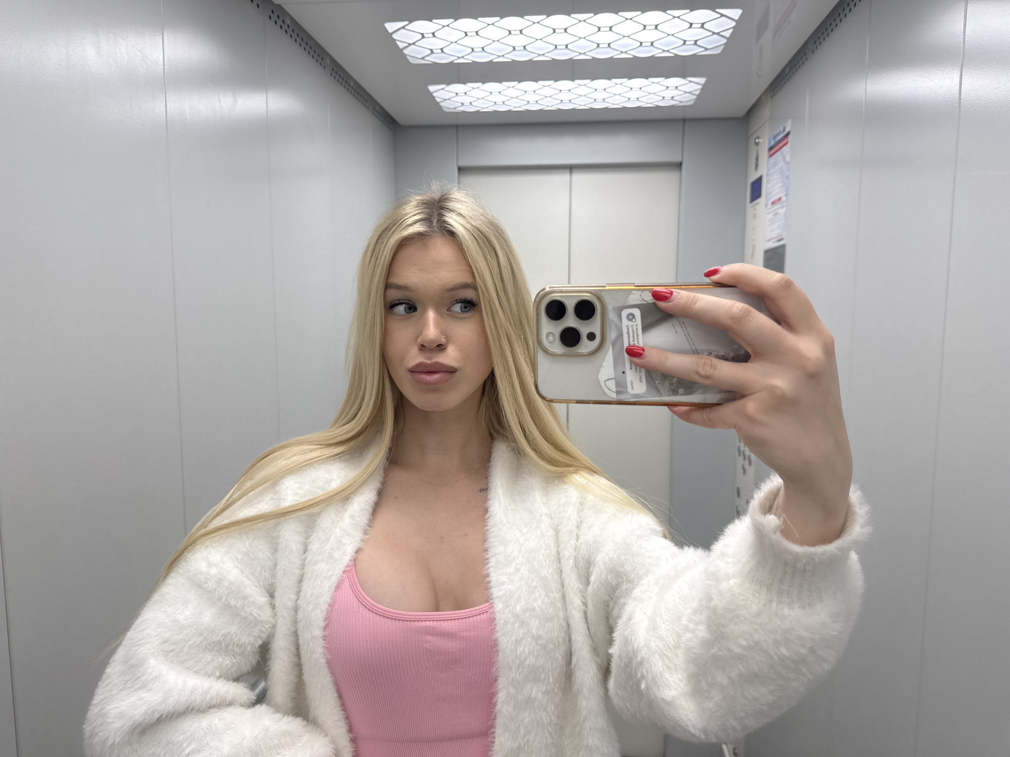 A young woman with blonde hair and full lips takes a mirror selfie in an elevator. She is wearing a pink tank top and a fluffy white cardigan. Her nails are painted red, and she is holding a smartphone in her right hand. The elevator walls are metallic gray, and the ceiling has two illuminated panels. The scene is indoors, with no identifiable context to suggest it is in Saint Petersburg, Russia.