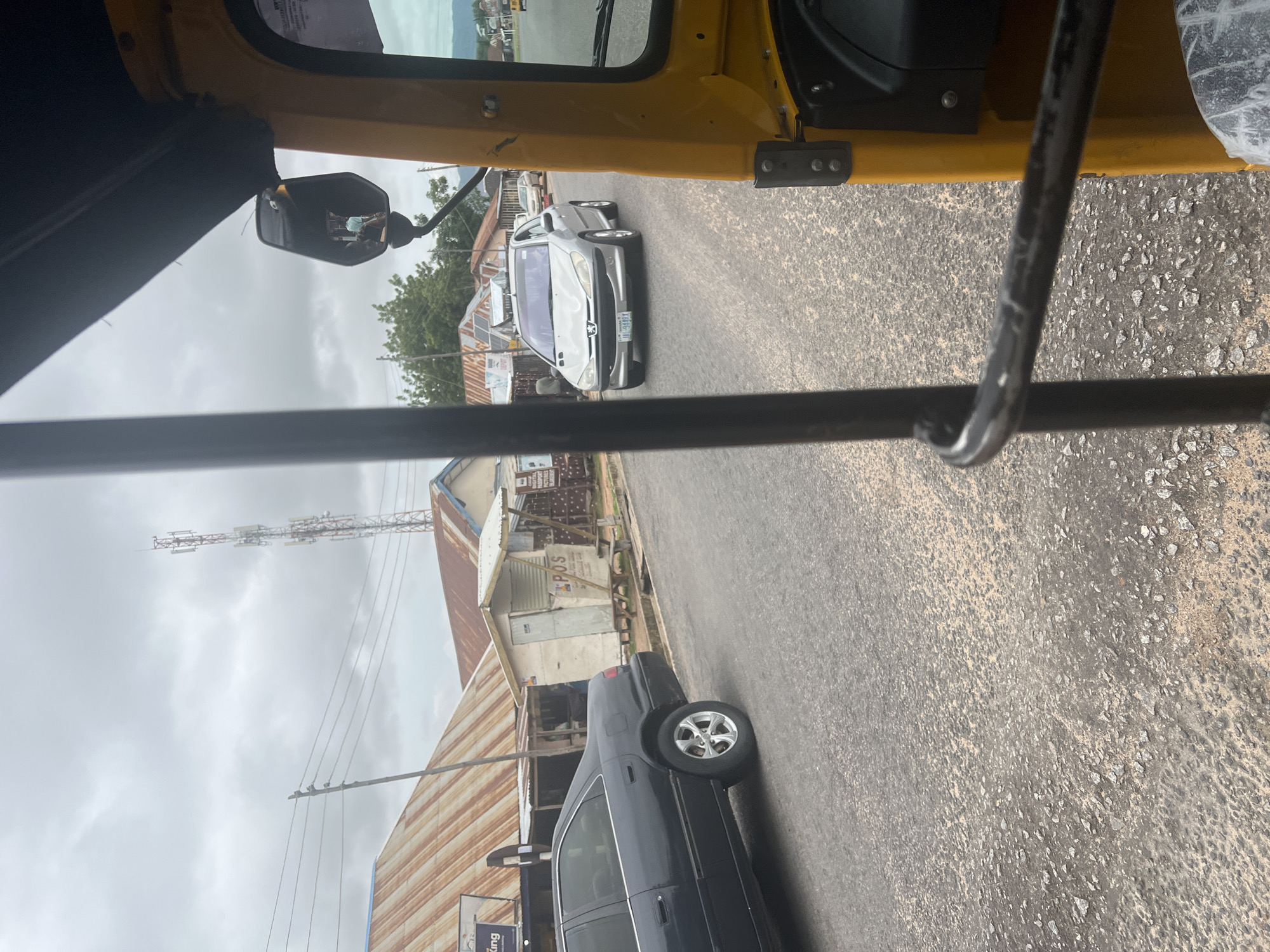 A low-angle, eye-level, point-of-view shot captures a street scene in Jalingo, Nigeria, from the perspective of someone inside a yellow auto-rickshaw. The foreground is dominated by the interior of the auto-rickshaw, with black metal bars framing the view. To the right, a portion of the yellow vehicle's interior is visible.
The street itself is paved with asphalt showing signs of wear and tear, with patches of lighter-colored gravel. On the left side of the street, a dark-colored sedan is parked or moving slowly. Beyond the car, a row of buildings with corrugated metal roofs, some appearing rusted, lines the street. One building has a white facade with the text "POS" clearly visible. Further down the street, another silver passenger car is in motion, its headlights on.
In the background, a tall, red and white telecommunications tower stands against a cloudy sky. Power lines are strung across the scene, supported by poles. Trees are visible in the distance. The overall impression is a typical streetscape in a Nigerian town, with a mix of residential or commercial structures, vehicles, and infrastructure. The lighting suggests it is daytime, possibly overcast.