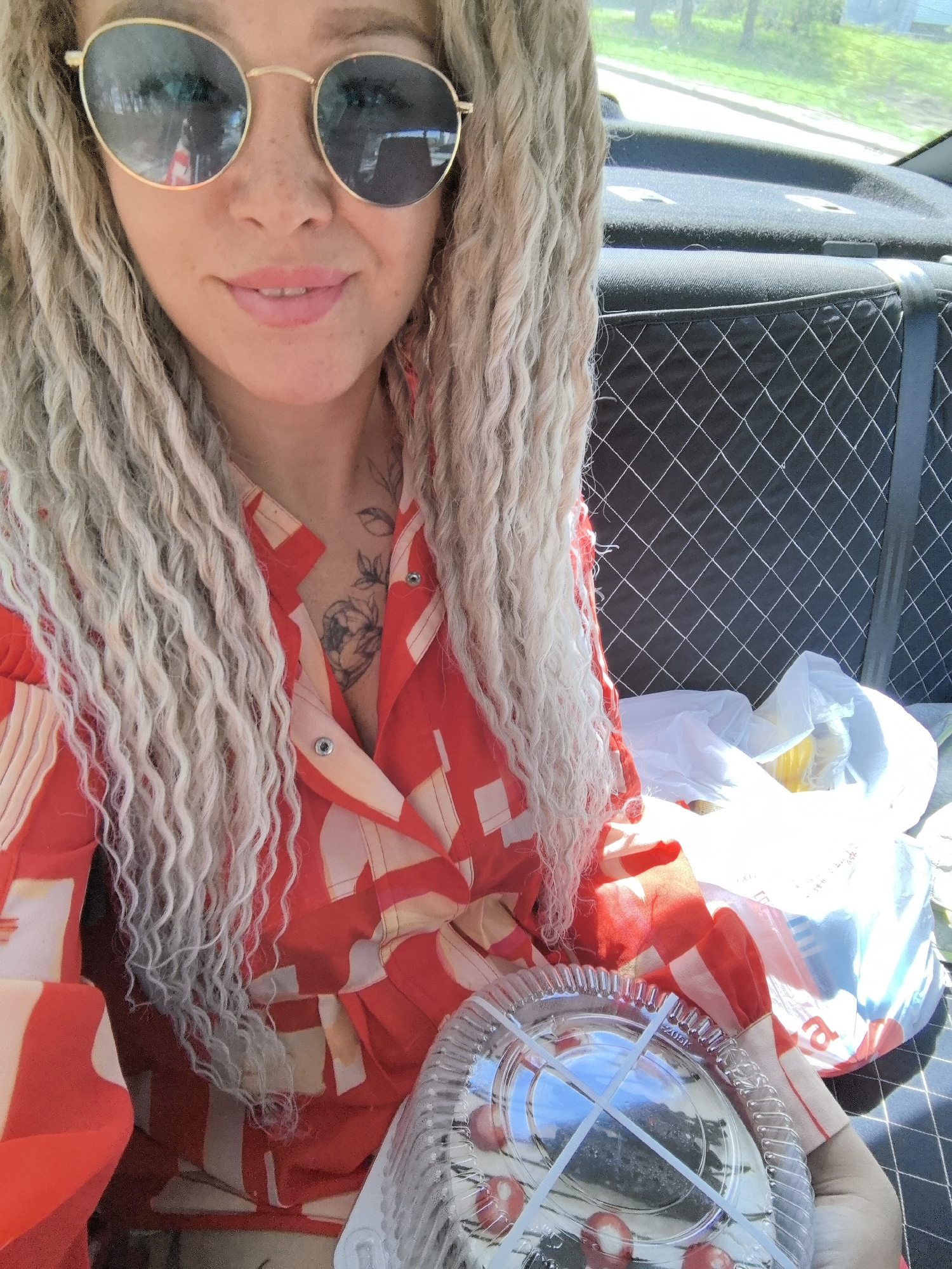 A woman with light blonde dreadlocks and wearing round gold-rimmed sunglasses is seated in a car, holding a plastic container with red cherries inside. She's wearing a red and beige geometric patterned shirt. A tattoo is visible on her chest. In the background are black car seats with a white diamond-shaped stitched pattern, along with shopping bags. The photo was taken in the city of Chaikovsky, Russia.