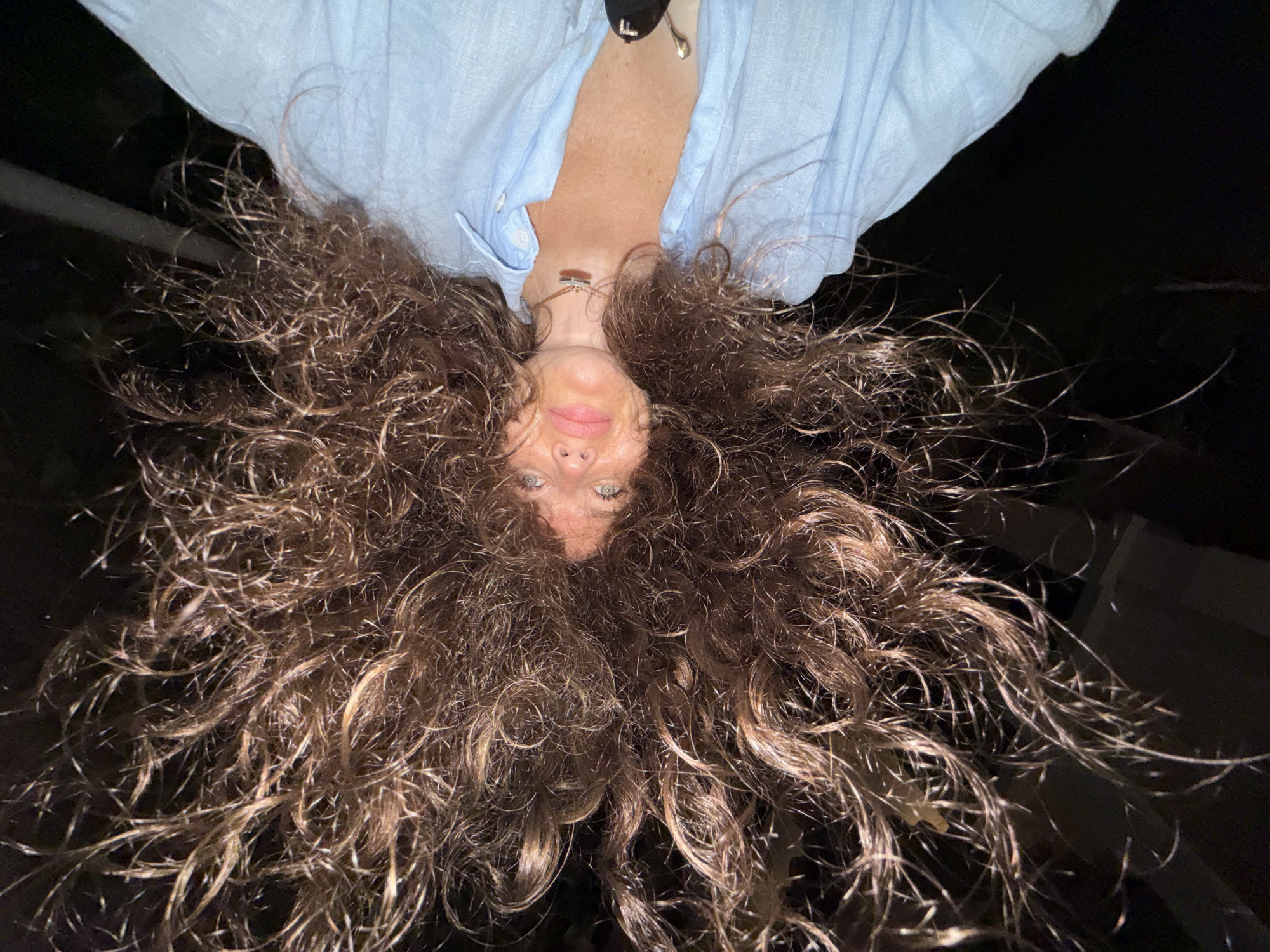 An upside-down, close-up shot of a woman with long, curly brown hair, her head tilted back, and her face obscured by her hair. She is wearing a light blue button-up shirt and a necklace. The background is dark and out of focus.