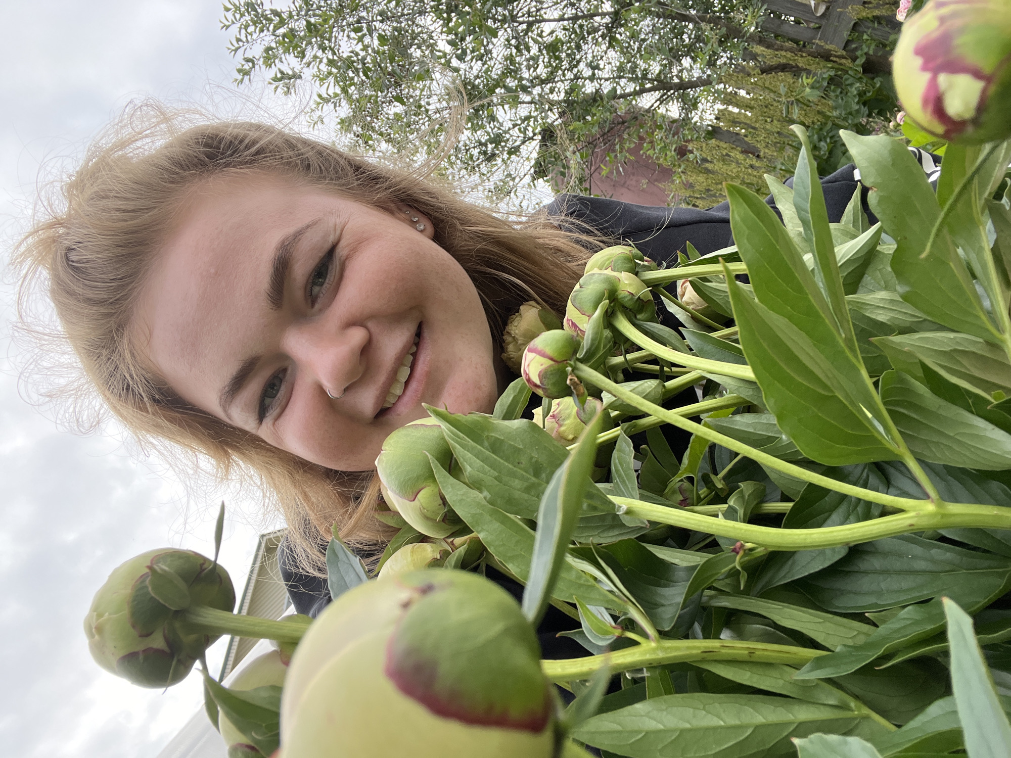 An overcast, outdoor, medium shot captures a young woman with blonde hair pulled back from her face, smiling broadly at the camera. She is positioned in the foreground, holding a large bouquet of unbloomed peonies. The peonies, predominantly green with hints of pink on the tips of their buds, are clustered together, their stems and lush green leaves filling the lower half of the frame. The woman is wearing a black hoodie. In the background, out of focus, is a wooden fence and foliage, suggesting a rural or suburban setting. The location is identified as Olkhovka, Russia.