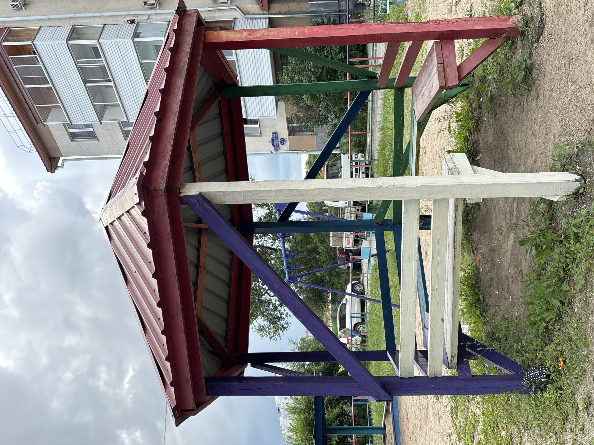 The image depicts a colorful, octagonal wooden gazebo situated in a playground area within the city of Blagoveshchensk, Russia. The gazebo's roof is a corrugated red metal, and its supporting posts are painted in various bright colors, including purple, green, and red. Benches are integrated into the structure, painted in white and green. In the background, a light-colored residential building with multiple balconies is visible. A street sign reading "ул. Дзержинского 41/2" (Dzerzhinsky Street 41/2) is affixed to the building, confirming the location. Other playground elements, such as a swing set and a sandpit, are partially visible. The ground is a mixture of sand and sparse vegetation. The sky is overcast, indicating a cloudy day. There are no people actively engaged in the scene, though a white SUV and a truck are parked in the middle distance.