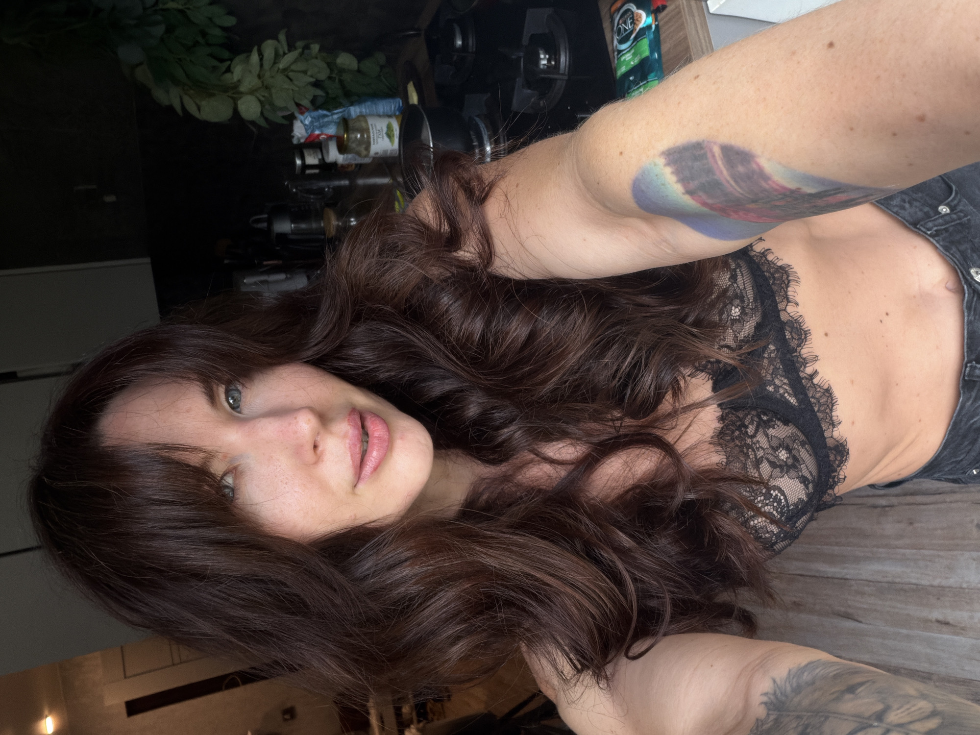 The image shows a woman taking a selfie in a kitchen. She has long, dark, wavy hair and is wearing a black lace bra and denim shorts. She has tattoos on her arms. The background shows kitchen cabinets, a stove, and various kitchen items. The image does not provide any information about the city of Perm, Russia.