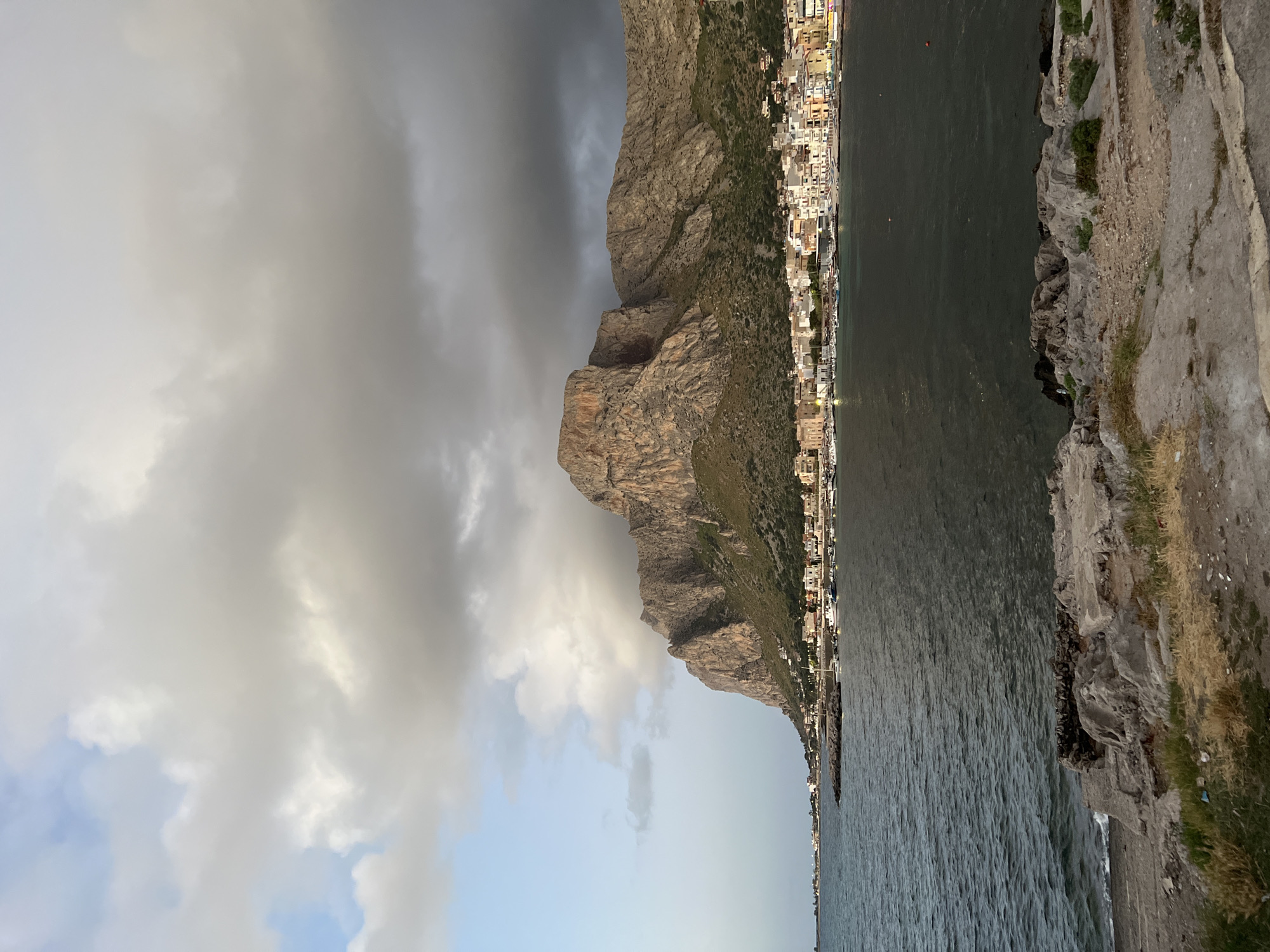 The image depicts a coastal view with a prominent mountain dominating the background, overlooking a town nestled at its base. The town, characterized by buildings with white and colored facades, lines the waterfront where numerous boats are docked in a harbor. The sea in the foreground is calm, with visible waves. The sky is partly cloudy, with a mix of dark and light clouds suggesting potential approaching weather. The terrain in the immediate foreground is rocky and somewhat uneven, with patches of dry grass. The scene appears to be captured during the golden hour or late afternoon, given the lighting on the mountain and town. The location is likely Castellammare del Golfo, a town situated on the northern coast of Sicily, which is part of the Metropolitan City of Palermo, Italy, known for its dramatic coastal landscapes and mountains.