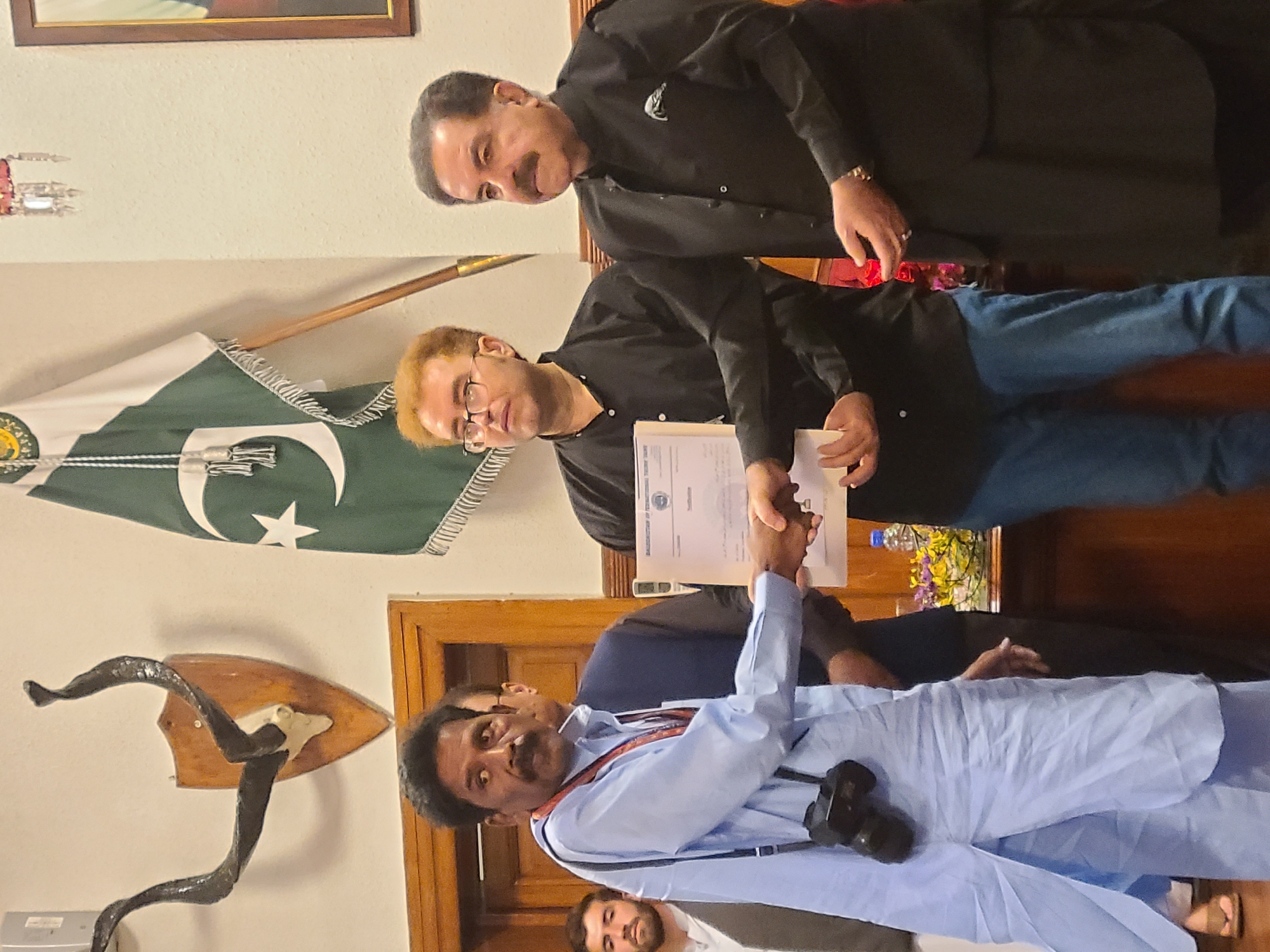 The image displays an indoor scene featuring four men engaged in a formal gathering. In the center, two men are shaking hands. The man on the right, wearing a black shirt, blue jeans, and spectacles, holds a document in his left hand while his right hand clasps the other man's. Partially visible text on this document includes "BALOCHISTAN BOARD OF TECHNICAL EDUCATION" and "TRAINING", indicating it is likely a certificate or official record. The man receiving the handshake wears a light blue shalwar kameez and has a camera hanging from a lanyard around his neck.
To the right of the central pair, another man in a black shirt and dark trousers with a mustache observes the interaction. At the bottom center, the head and shoulders of a fourth man with dark hair and a beard are visible.
The background features a Pakistan national flag, characterized by its green field, white vertical stripe, and white crescent and star, hanging prominently on the left wall. Above the flag, the lower edge of a framed picture is seen. Below the flag, a wall-mounted decorative item resembling horns or antlers is displayed. To the right, a vase containing flowers and a water bottle are placed on a surface, next to a black sofa. The wooden doorframe or paneling is also visible on the left. The scene suggests an official ceremony or presentation, consistent with a formal setting in Quetta City Tehsil, Pakistan. The men appear engaged and focused on the exchange.