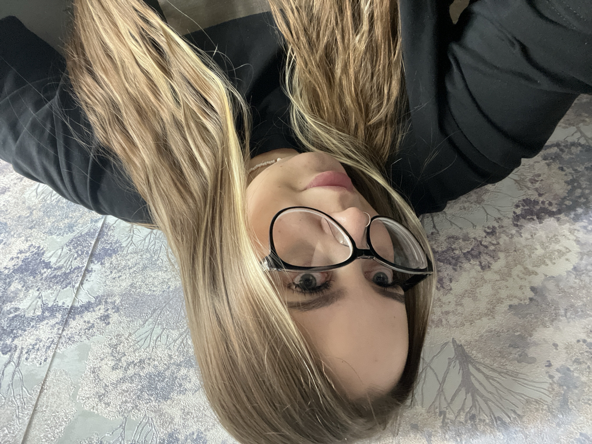 A selfie of a fair-skinned person with blonde hair and black-framed glasses is presented in a rotated perspective. The person is wearing a dark-colored garment. The background is a textured surface, possibly a bedspread or wallpaper, with a tree-like pattern in shades of gray and white. No evidence suggests the location is in Novopokrovskaya, Russia.