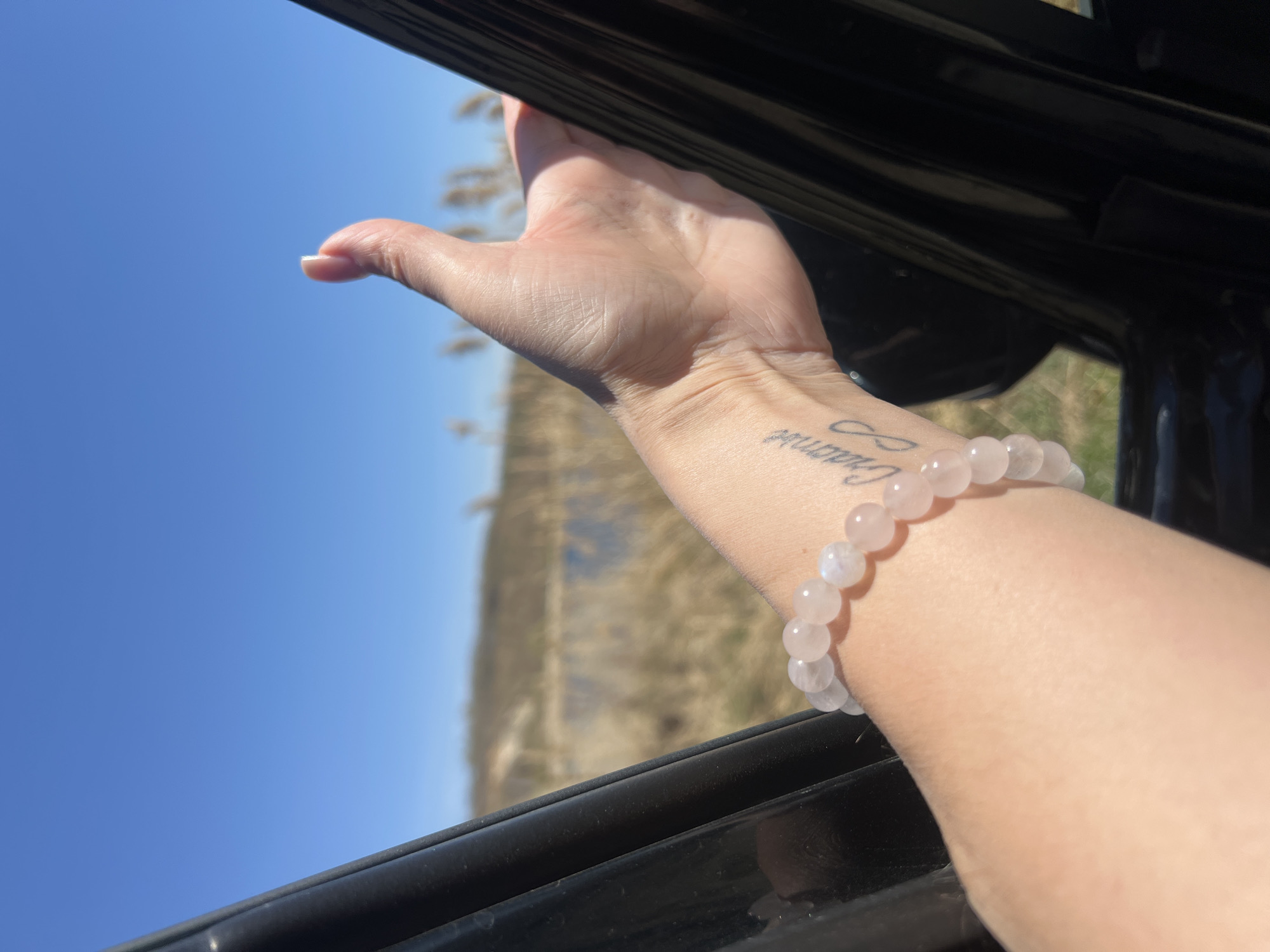 A close-up shot captures a person's arm extending from a car window, with the blue sky and blurred landscape in the background. The arm features a pale pink beaded bracelet and a tattoo, appearing near the wrist. The car window frame is black and glossy. The setting is likely outdoors, with natural light illuminating the scene. The location is Unknown, Unknown.