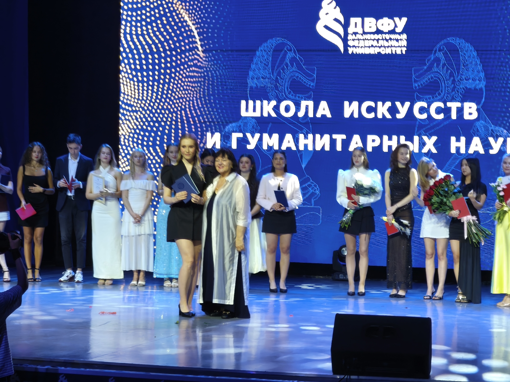The image shows a group of young people and some adults standing on a stage in front of a large screen displaying text and graphics. The screen shows the logo of "ДВФУ" (Far Eastern Federal University) and the words "ШКОЛА ИСКУССТВ И ГУМАНИТАРНЫХ НАУК" (School of Arts and Humanities). The people are dressed in formal or semi-formal attire, and some are holding certificates or bouquets of flowers. They appear to be participating in an award ceremony or a graduation event. The stage is illuminated by stage lights, and the floor is reflective, showing the lights and the audience, partially visible at the bottom. The event is likely taking place at the Far Eastern Federal University campus on Russky Island, in Vladivostok, Russia, given the university's name displayed on the screen.