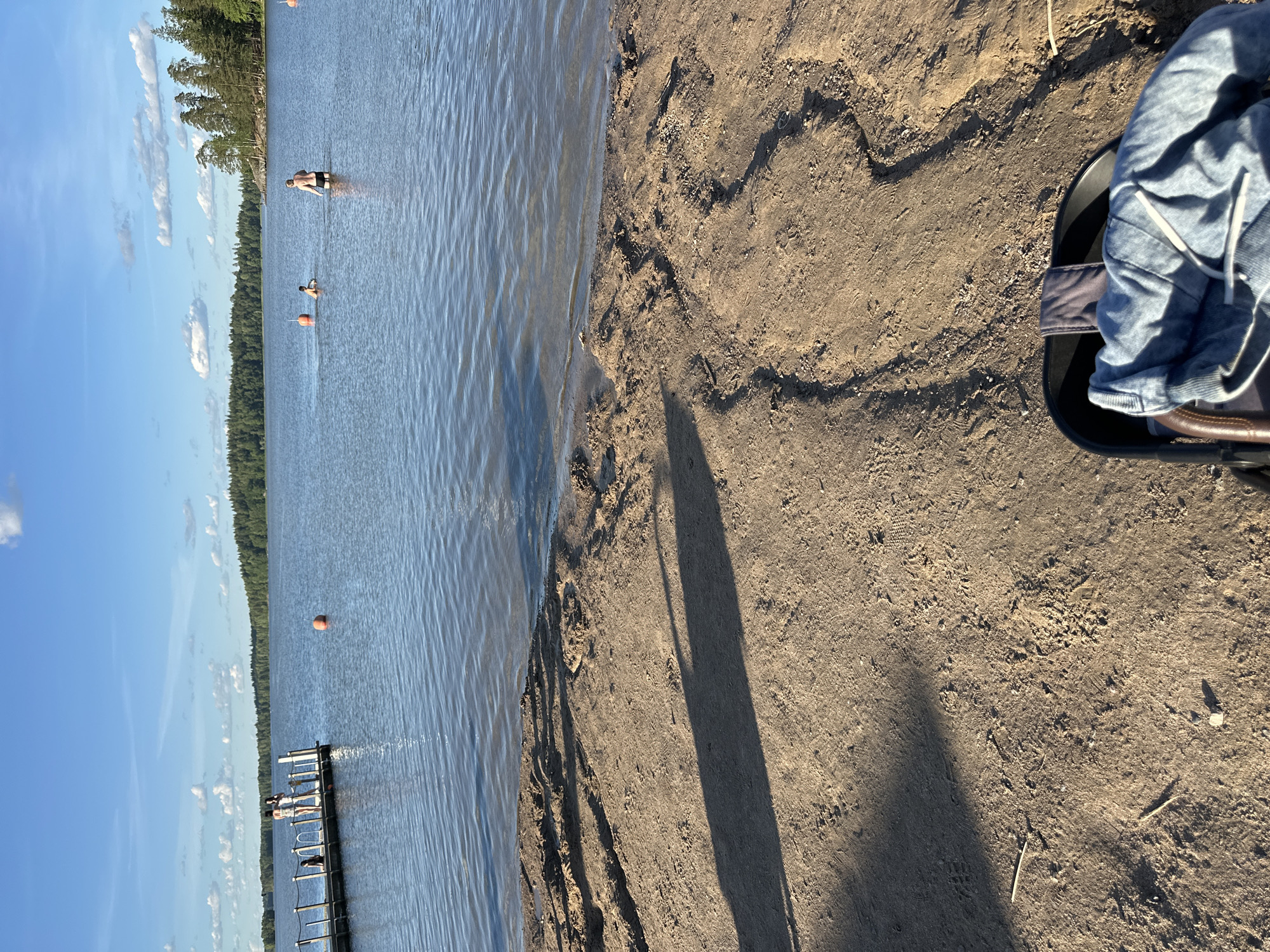 An outdoor, daytime, eye-level shot depicts a beach and a body of water. The foreground features a sandy beach with tire tracks and shadows from an unseen object, possibly a stroller, with clothing on it. In the midground, the water is calm with a few buoys and ripples. Several people are visible in the water and on a wooden dock. The background shows a tree-lined shore with a bright blue sky dotted with white clouds.