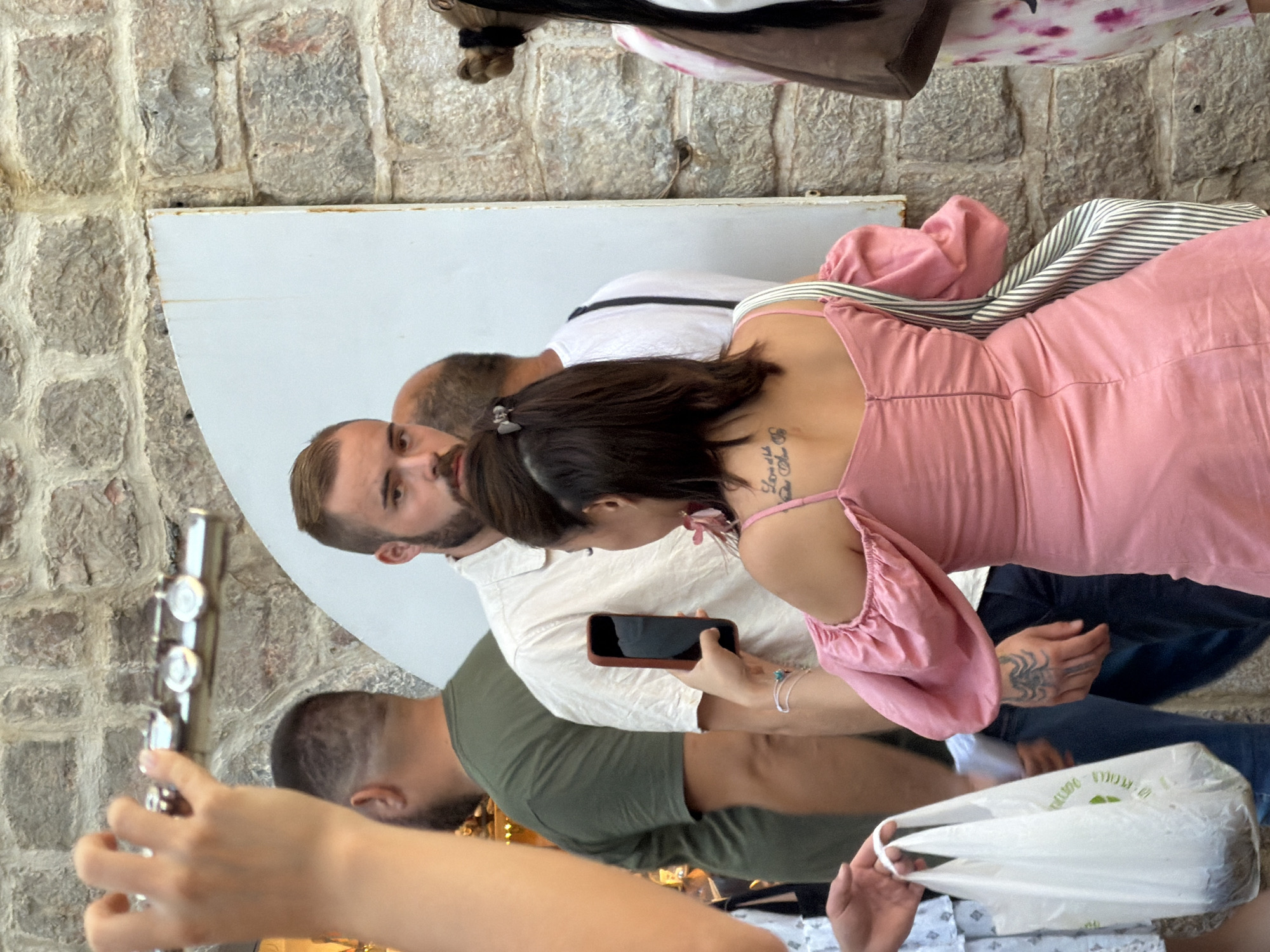 In Budva, Montenegro, a group of people are gathered near a stone wall. A woman in a pink dress is holding a smartphone to take a photo. She has a tattoo on her arm. Another person in a green shirt is standing behind her. In the background, a man with a beard looks towards the camera. A white plastic bag is held by a person on the left side of the frame. The scene appears to be outdoors, possibly in a pedestrian area or street.