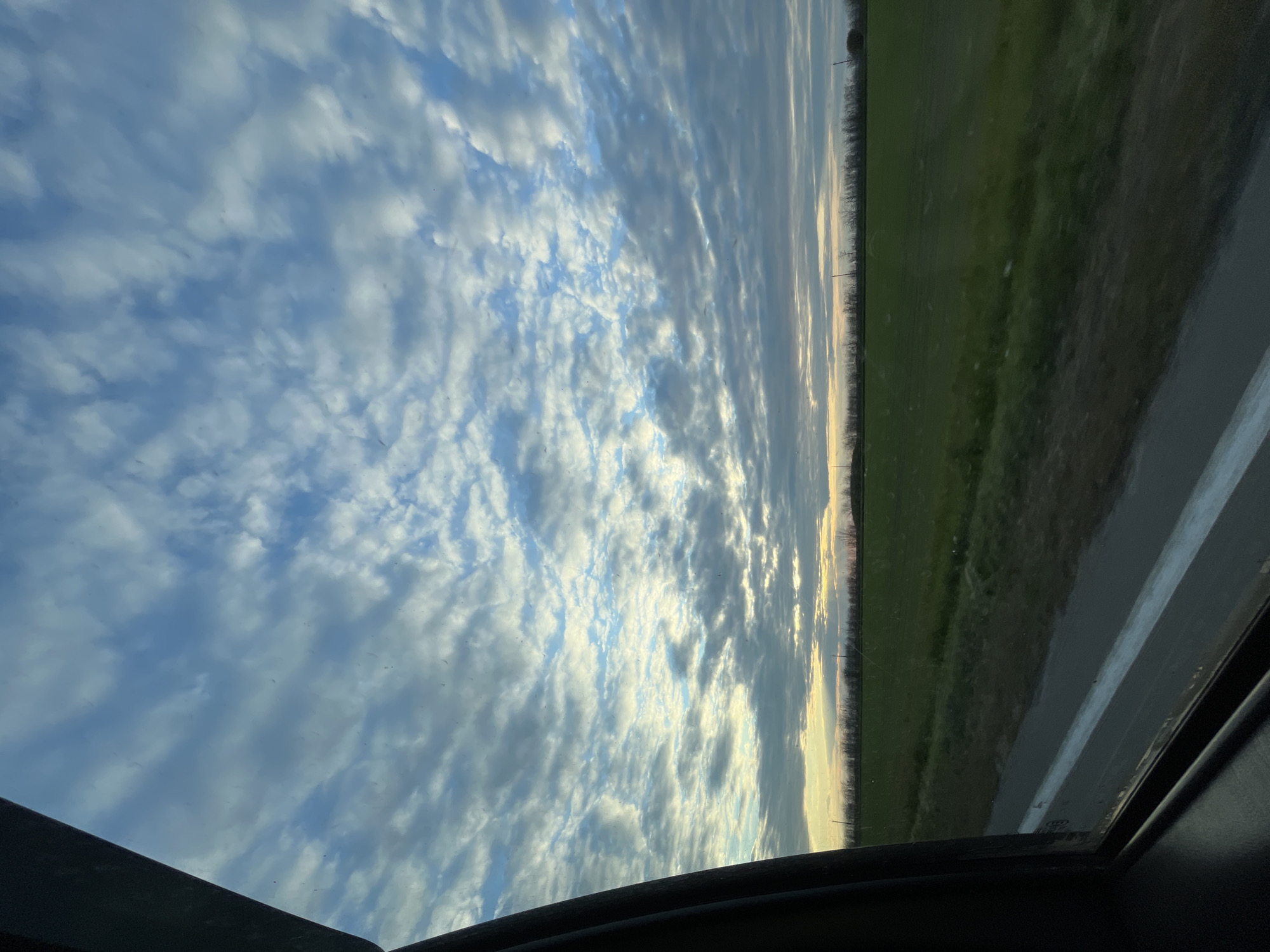 The image shows a view from inside a vehicle, likely a car, looking out the driver's side window. The landscape includes a field and a road. The sky is predominantly blue with scattered clouds. Based on the image content alone, it is not possible to confirm or deny the location of the photograph in Novomargaritovo, Russia.