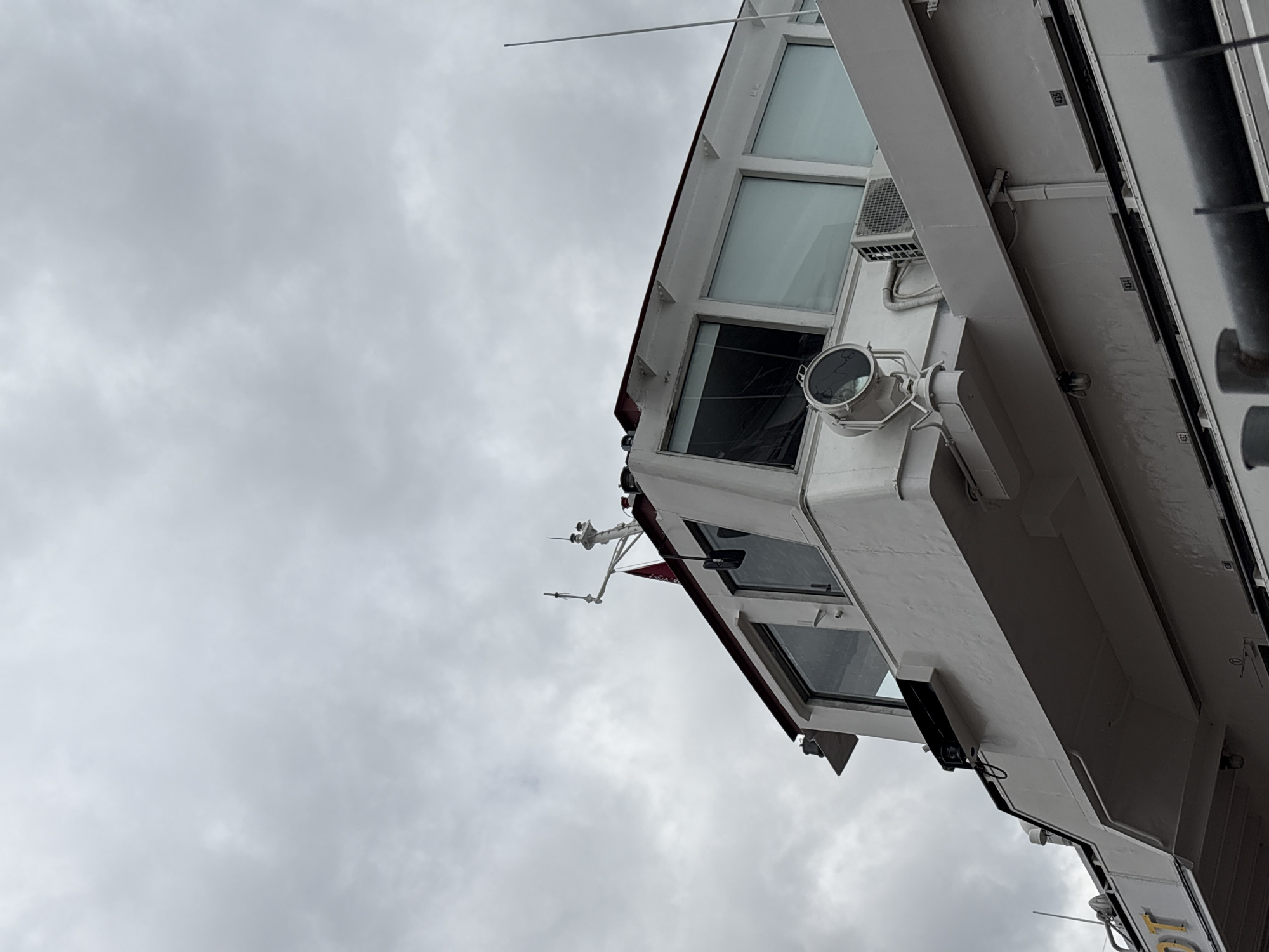 This is a low-angle, medium shot of the bridge of a white ferry boat under a cloudy sky. The ferry appears to be docked or anchored. The bridge has large windows and a spotlight mounted on the exterior. A red flag is visible on a mast. The boat has the word "IDOT" visible in yellow lettering on its side. The location is identified as Shushary, Russia.