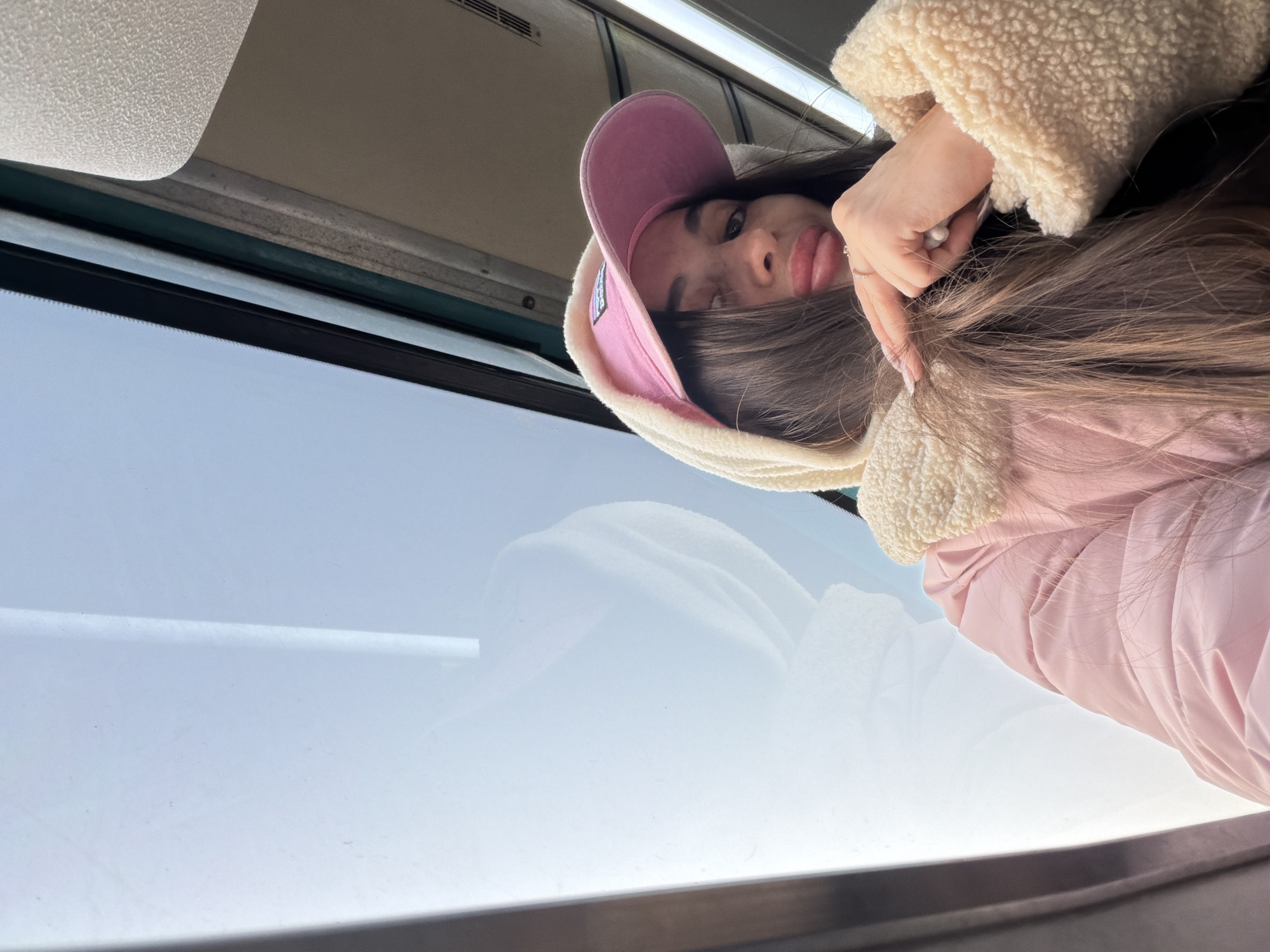 A woman wearing a pink hat and a beige, sherpa-lined jacket is seen in the image. She is positioned close to a window. Based on the architectural details seen through the window, the location is likely Saint Petersburg, Russia, but this is not definitive without further context or visual cues.