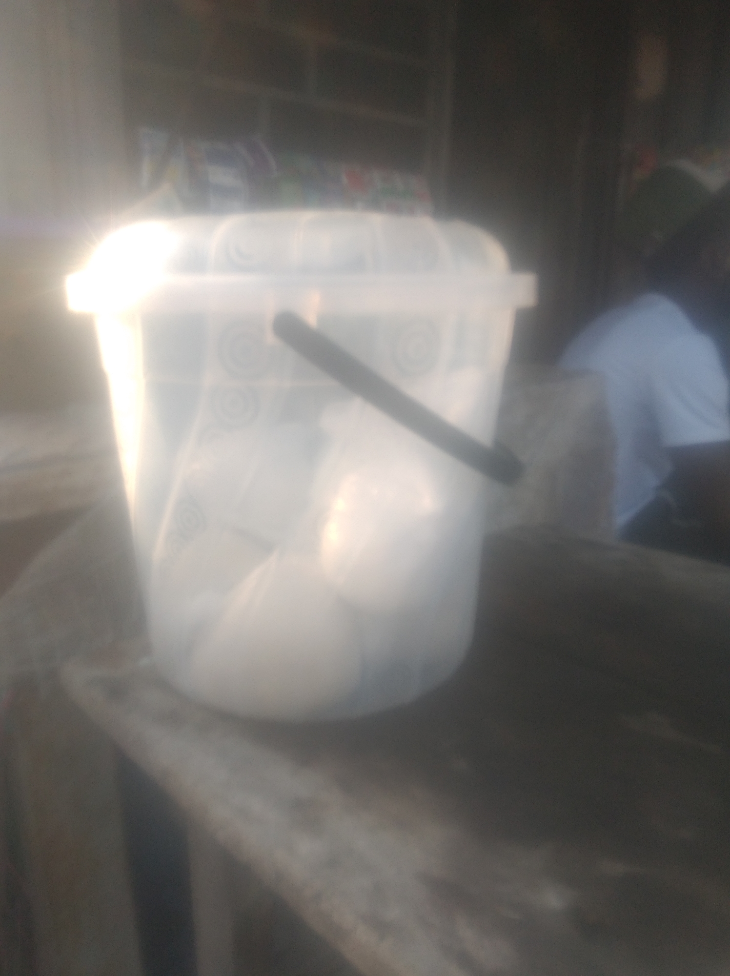 In the city of Jalingo, Nigeria, an out-of-focus image captures a partially translucent white plastic bucket on a rough wooden surface. The bucket appears to contain several white, opaque objects, possibly bags. A dark handle extends from the bucket's rim. To the right, a person wearing a light blue shirt is visible, their face obscured by blur and shadow. The background is dark and indistinct, suggesting an indoor or shaded outdoor setting. The overall lighting is bright, with a strong glare from the left side of the image.