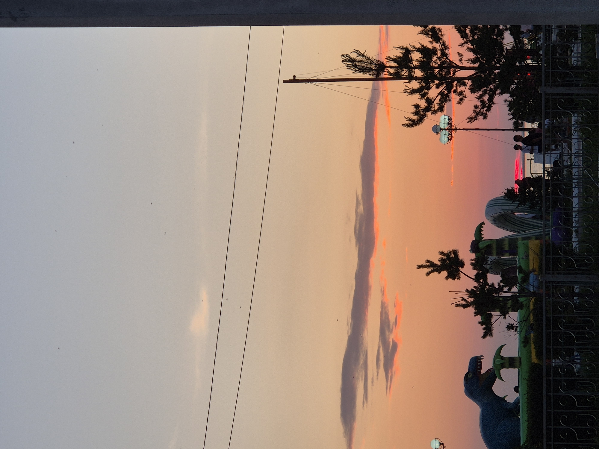 The image shows an outdoor scene at dusk, likely in Kokshetau, Kazakhstan. The sky displays a gradient of colors from light blue at the top to orange and pink hues near the horizon, indicating sunset. Several dark clouds are visible, some tinged with pink from the setting sun. Power lines stretch horizontally across the upper portion of the sky.
In the foreground, silhouetted against the sunset, is a fenced area containing inflatable structures, possibly part of a children's play area or amusement park. A large blue dinosaur inflatable is prominent on the left. To the right, a tall evergreen tree stands near a lamppost with multiple globes. Several people are visible in silhouette near the water's edge in the background, suggesting a waterfront location. A thick, vertical concrete pillar frames the right side of the image. The overall atmosphere is tranquil, capturing the end of the day.