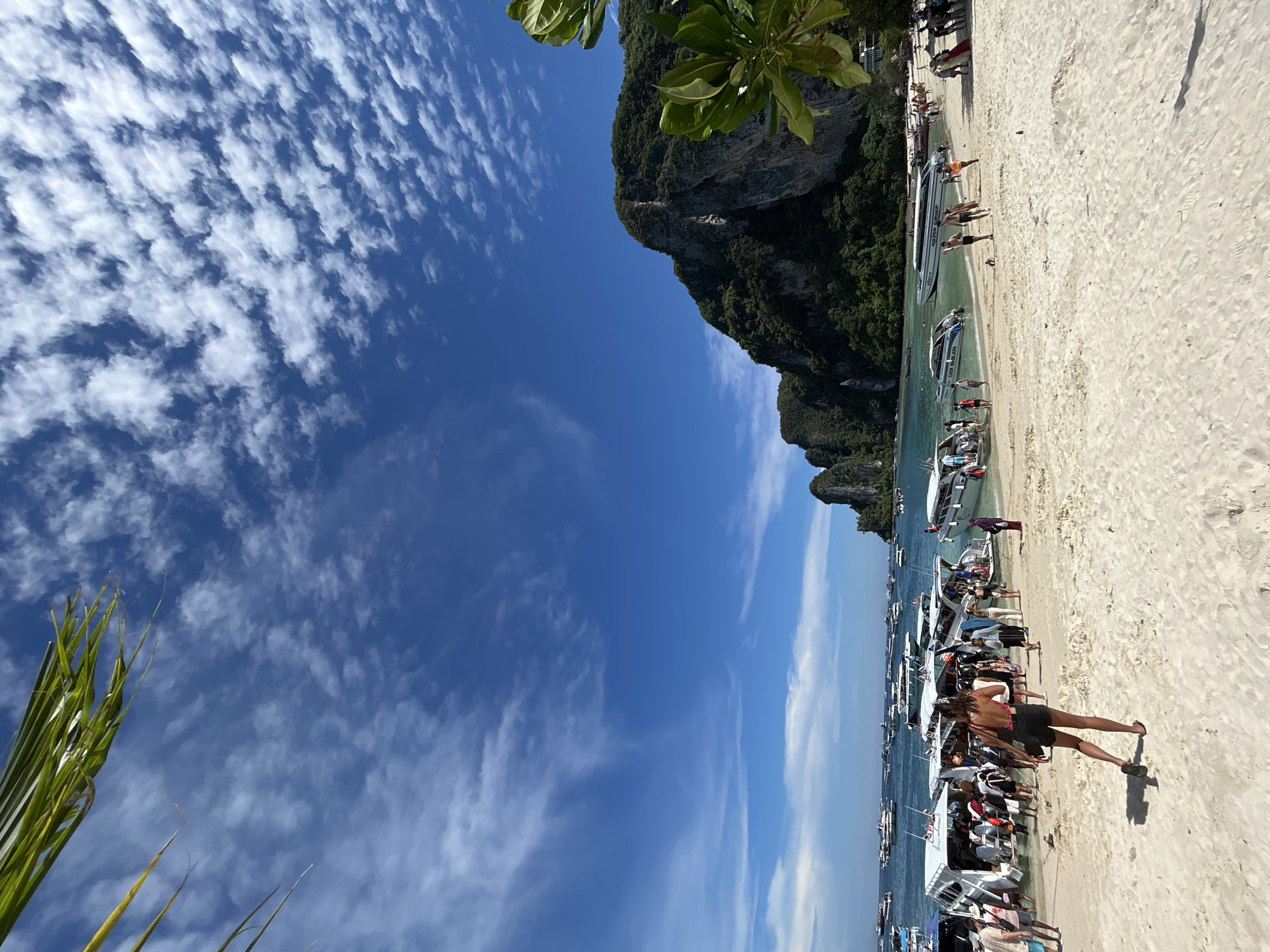 This image depicts a busy beach scene in Ao Nang, Thailand, likely at a departure point for island tours. The foreground is a white sand beach with several people milling about. A woman in black shorts and a red and white patterned top walks away from the camera towards the water. In the midground, numerous speedboats are docked or anchored near the shore, many of them carrying passengers. The boats are white with blue accents and some have identifying text on their sides. Tourists can be seen boarding and disembarking these boats. The background features a lush, green, rocky cliff face rising out of the turquoise water. The sky is a vibrant blue with scattered white, wispy clouds and a hint of palm fronds in the upper left corner. The scene is bathed in bright sunlight, suggesting a warm, tropical day.