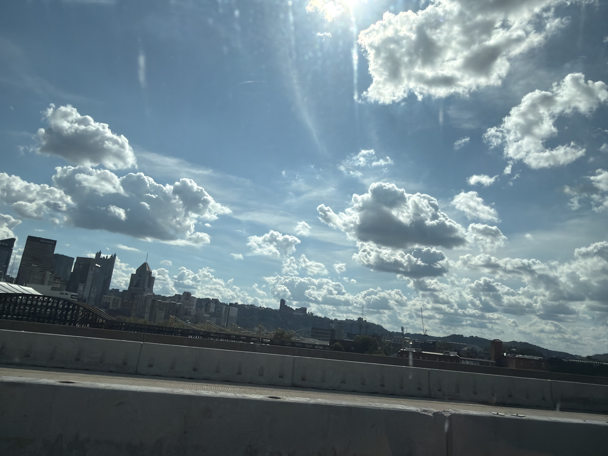 The image shows a daytime view of the Pittsburgh skyline, with the city's distinctive architecture visible in the distance. The foreground consists of a concrete highway barrier, suggesting the photograph was taken from a vehicle traveling on an elevated roadway or bridge. Above the city, a vast expanse of blue sky is dotted with numerous white, fluffy clouds. The sun is prominently visible in the upper right quadrant of the image, casting bright rays and creating lens flare effects. The clouds are varied in size and density, with some appearing more shadowed and dramatic than others. In the mid-ground, the city's buildings, including skyscrapers and other structures, rise against the backdrop of rolling hills in the far distance. A bridge with a distinctive lattice-work structure is partially visible in front of the cityscape. The overall scene depicts a metropolitan area under a dynamic, cloud-filled sky.