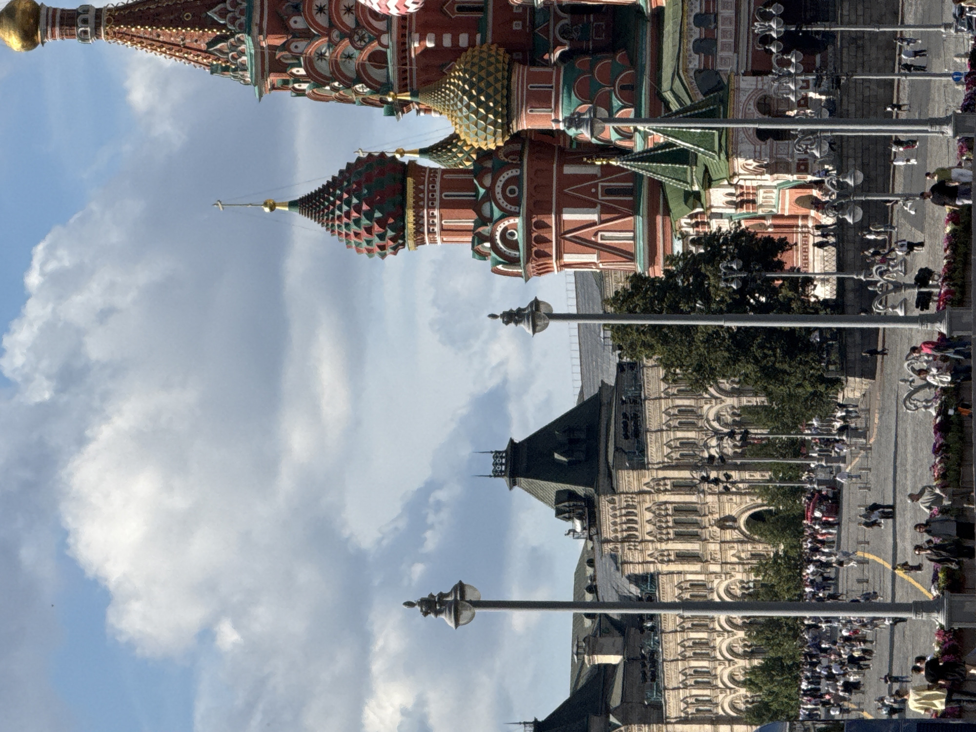 This is a vertical, eye-level, full shot of St. Basil's Cathedral and the GUM department store on Red Square in Moscow, Russia. The sky is a bright blue with scattered white clouds. The foreground features a cobblestone square filled with people walking, standing, and mingling. Several lampposts with multiple lights are visible. In the middle ground, the ornate, multi-colored domes and spires of St. Basil's Cathedral dominate the right side of the frame, with its distinctive red and green patterns and golden accents. To the left of the cathedral is the long, impressive facade of the GUM department store, a light-colored building with arched windows and a dark, pitched roof. Trees are interspersed throughout the scene. The overall atmosphere appears to be that of a busy, sunny day on a significant historical and tourist site.