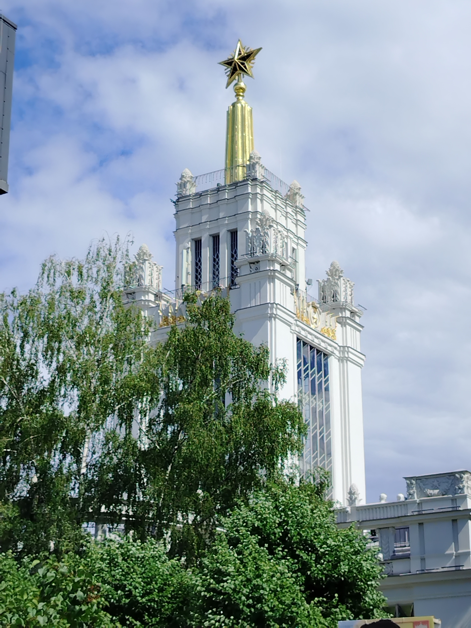 The image shows the towering spire of the main pavilion of the VDNKh (Exhibition of Achievements of National Economy) in Moscow, Russia, topped by a gilded star. The architecture is white with gold accents, characteristic of Soviet neoclassical style. Lush green trees with delicate leaves, likely birch, partially obscure the lower portion of the pavilion, creating a contrast between the man-made structure and the natural elements. The sky is blue with scattered white clouds, suggesting pleasant weather. The scene is a static, daytime exterior shot focusing on a prominent landmark within the VDNKh complex. There are no people or immediate actions depicted.