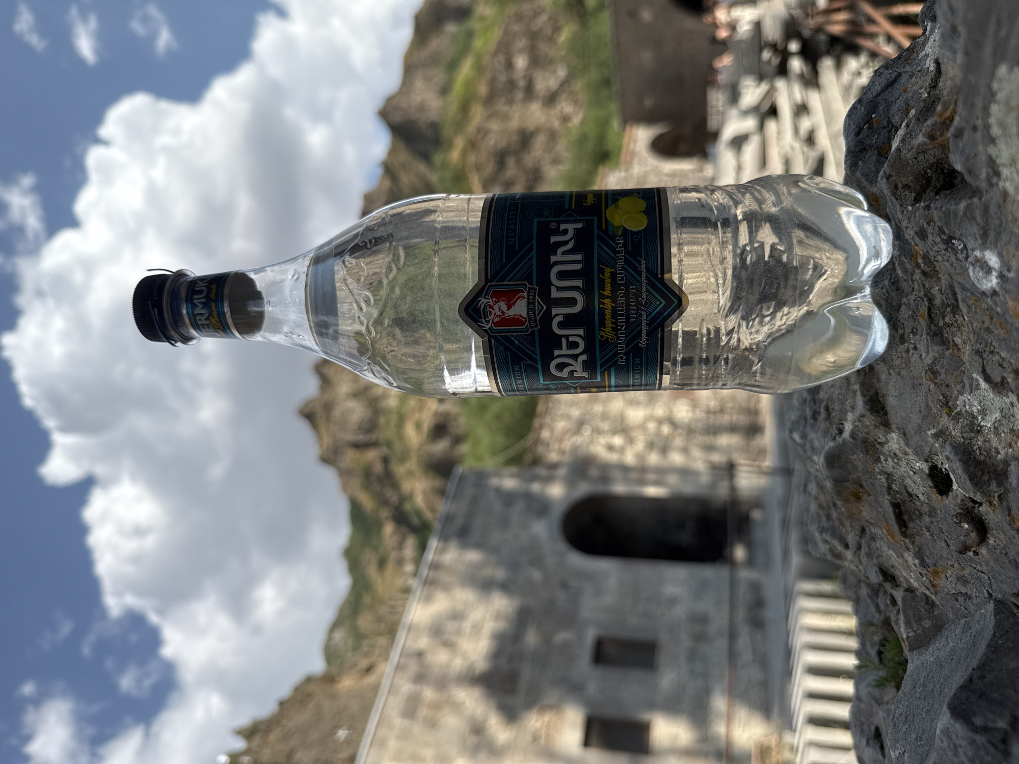A clear plastic bottle of Jermuk brand mineral water stands on a rough, grey rock in the foreground. The bottle has a dark blue and teal label with Armenian script, a deer emblem, and an illustration of lemons. The background is out of focus and shows stone structures, hills, and a cloudy blue sky. The stone structures appear to be ancient or historical, possibly part of a monastery or ruin, and are situated in a mountainous landscape. The overall scene suggests an outdoor setting in a place with historical significance, possibly in Goght, Armenia, given the mention of Jermuk water, which is famously sourced from Jermuk city. There are no visible people in the immediate foreground, but blurry figures can be seen in the background.