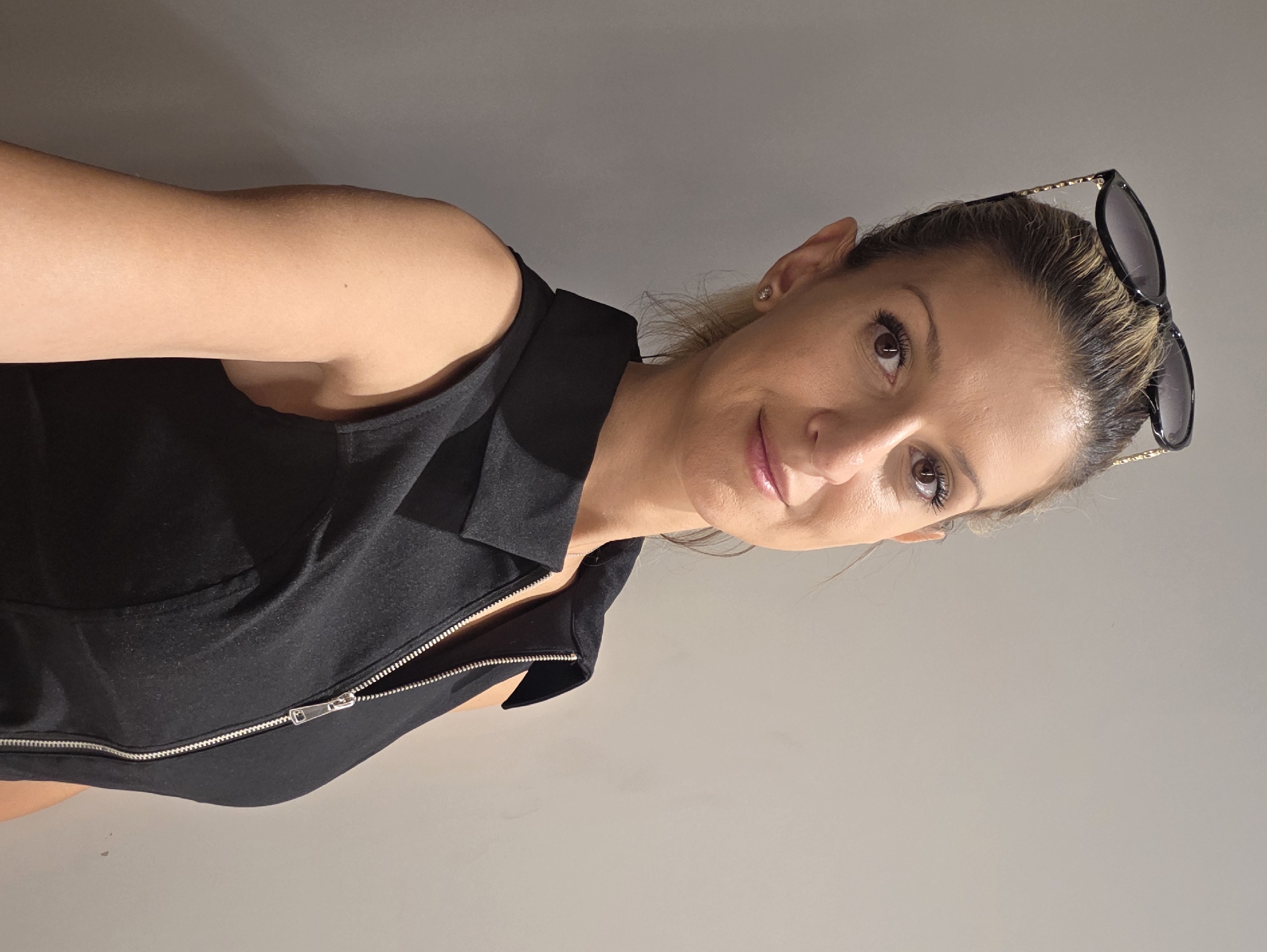 A woman is posing for a selfie against a plain grey wall. She is wearing a black sleeveless dress with a zipper down the front and sunglasses perched on top of her head. The lighting suggests it might be indoors. There is no indication of the location being Munich, Germany.