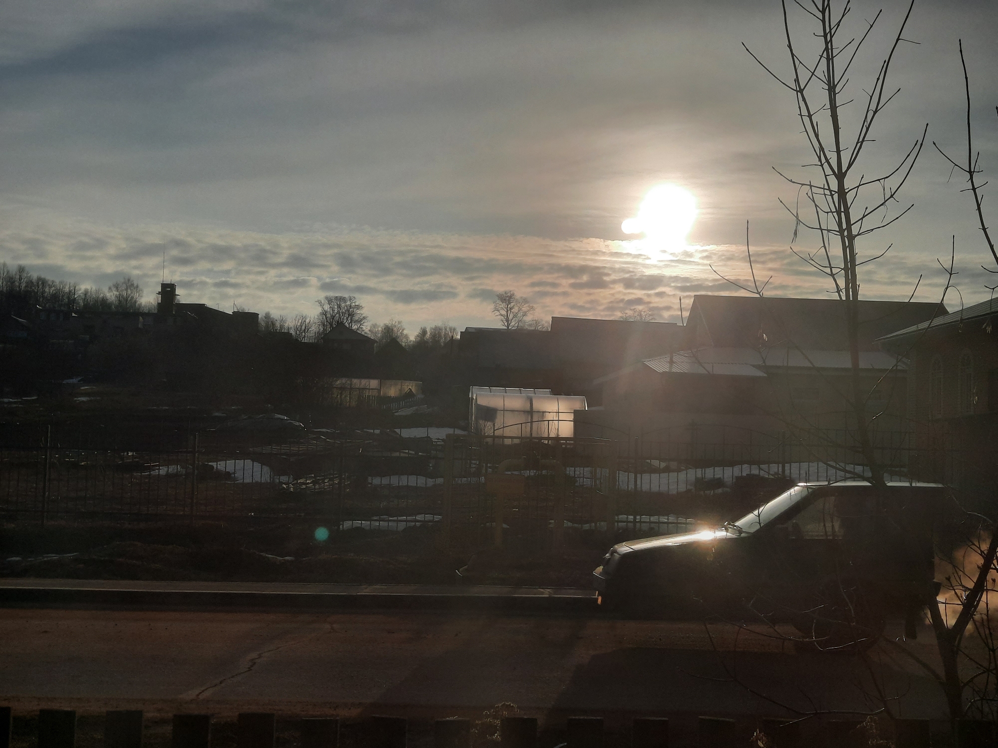 The image captures a residential scene under a bright, partially cloudy sky with the sun as a dominant light source. Buildings are visible in the background, along with what appears to be a greenhouse. A dark car is parked on the roadside, with sunlight reflecting off its surface. Bare trees are positioned in the foreground, adding to the silhouette effect created by the backlighting. The location appears to be on the outskirts of a town or city, as indicated by the mix of residential buildings, open spaces, and sparse vegetation. The presence of residual snow suggests a location with a cold climate, while the low angle of the sun may suggest it's either early morning or late afternoon.