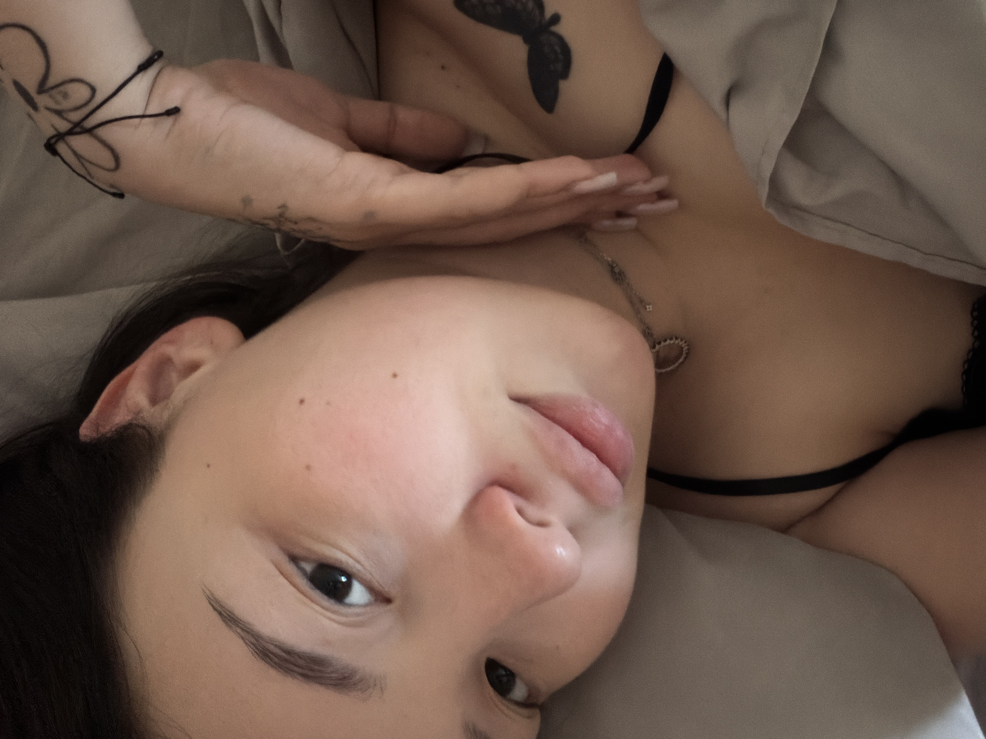 The media file is a close-up, overhead shot of a person lying in bed, seemingly in the morning. The person's face is tilted towards the camera, with their eyes looking directly at the viewer. They have dark hair and fair skin, with a smattering of freckles across their nose and cheeks. A delicate necklace with a heart-shaped pendant rests on their collarbone.
The person is wearing a black bra, with a glimpse of underwear visible. Their left arm is raised and positioned near their face, with a black string bracelet on their wrist and a tattoo of a flower. Another tattoo of a butterfly is visible on their chest. The setting appears to be a bedroom, with soft, neutral-colored bedding. The lighting is soft and diffused, suggesting it's either early morning or overcast. The overall mood is intimate and relaxed. There is no visible text in the image.