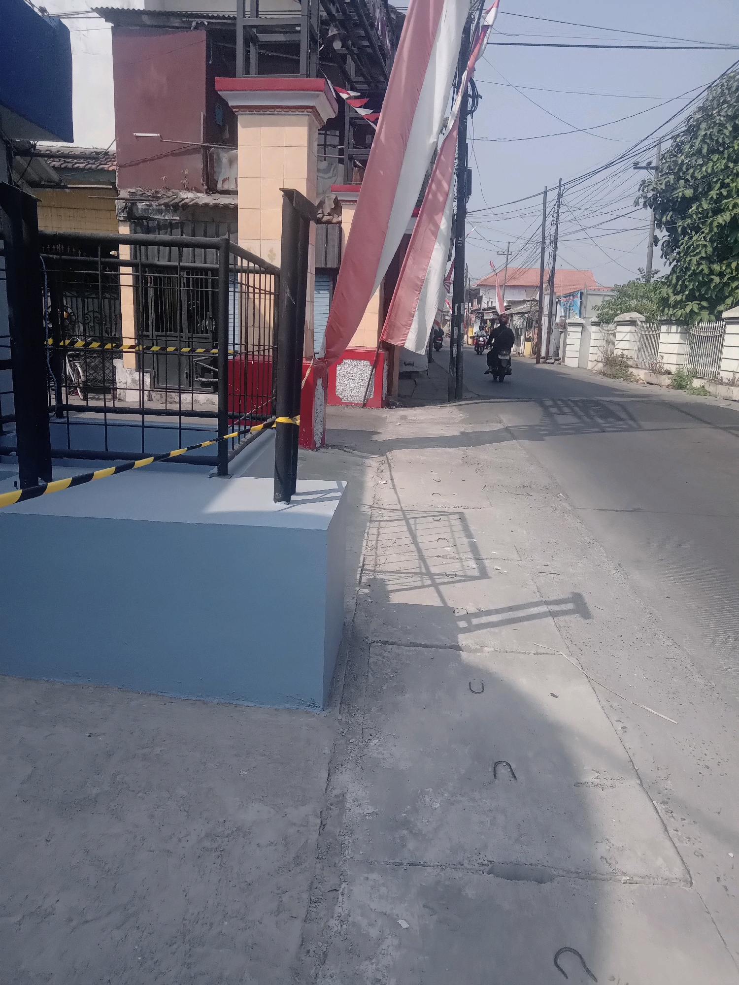 This is a street-level, medium-wide shot of a road in Wage, Indonesia, under a clear sky. The view is dominated by a grey concrete sidewalk in the foreground, with a freshly painted, light blue raised concrete structure to the left, marked with yellow and black striped tape. Beyond this structure is a black metal fence, with a bicycle visible behind it.
Further down the street, there are buildings with red and yellow facades, and a prominent pole adorned with Indonesian flags. Several people on motorcycles are traveling down the street, casting shadows on the asphalt. The road is lined with power lines strung across multiple poles. To the right, a white decorative fence separates the sidewalk from a dense green tree. The overall impression is of a residential street during the day.