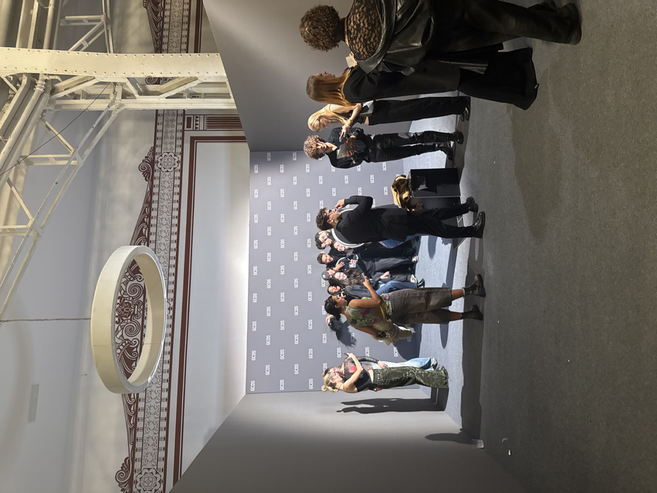 The image depicts an indoor gathering, likely an event for the Italian fashion brand GCDS, consistent with events typically hosted in Milan, Italy. The scene features a branded photo backdrop and several individuals.
**Environment:** The setting is a large room with a high white ceiling. Ornate reddish-brown patterns decorate the upper portion of the permanent walls. White industrial-style metal beams are visible in the upper right. A large, white, circular light fixture is suspended from the ceiling in the upper left. The floor is covered with a dark gray carpet. Temporary gray partition walls create a corner, with the primary backdrop wall featuring a repeating "GCDS" logo pattern against a lighter gray background.
**People:** Approximately 15-20 individuals are present, predominantly young adults, dressed in contemporary attire.
* **Left Side:** A blonde female in a dark patterned top and wide-leg green pants is taking a photo with a smartphone, facing the GCDS backdrop. Another person is partially visible behind her.
* **Center-Left:** A female with dark hair, wearing a multi-colored top and long brown shorts, stands looking down at a smartphone.
* **Center:** A cluster of 10-12 people is gathered in front of the GCDS backdrop, many smiling and looking towards the left, consistent with posing for a photo. Several are holding smartphones.
* **Mid-Ground Right:** A male in a black suit and glasses is positioned facing right, with his right hand near his mouth. A light brown jacket is draped over a black cube-shaped object beside him.
* **Right Side:** A tall male with curly hair, dressed entirely in black, is looking at a device while standing next to a blonde female who is also holding a device. Further right, two additional females are present; one in a dark long dress or skirt, and another seen from behind, wearing a black lace-detailed top and a black jacket draped over her shoulders.
**Actions and Interactions:** Individuals are engaged in social interaction, posing for photographs, and using their mobile phones. The overall atmosphere is lively and casual.