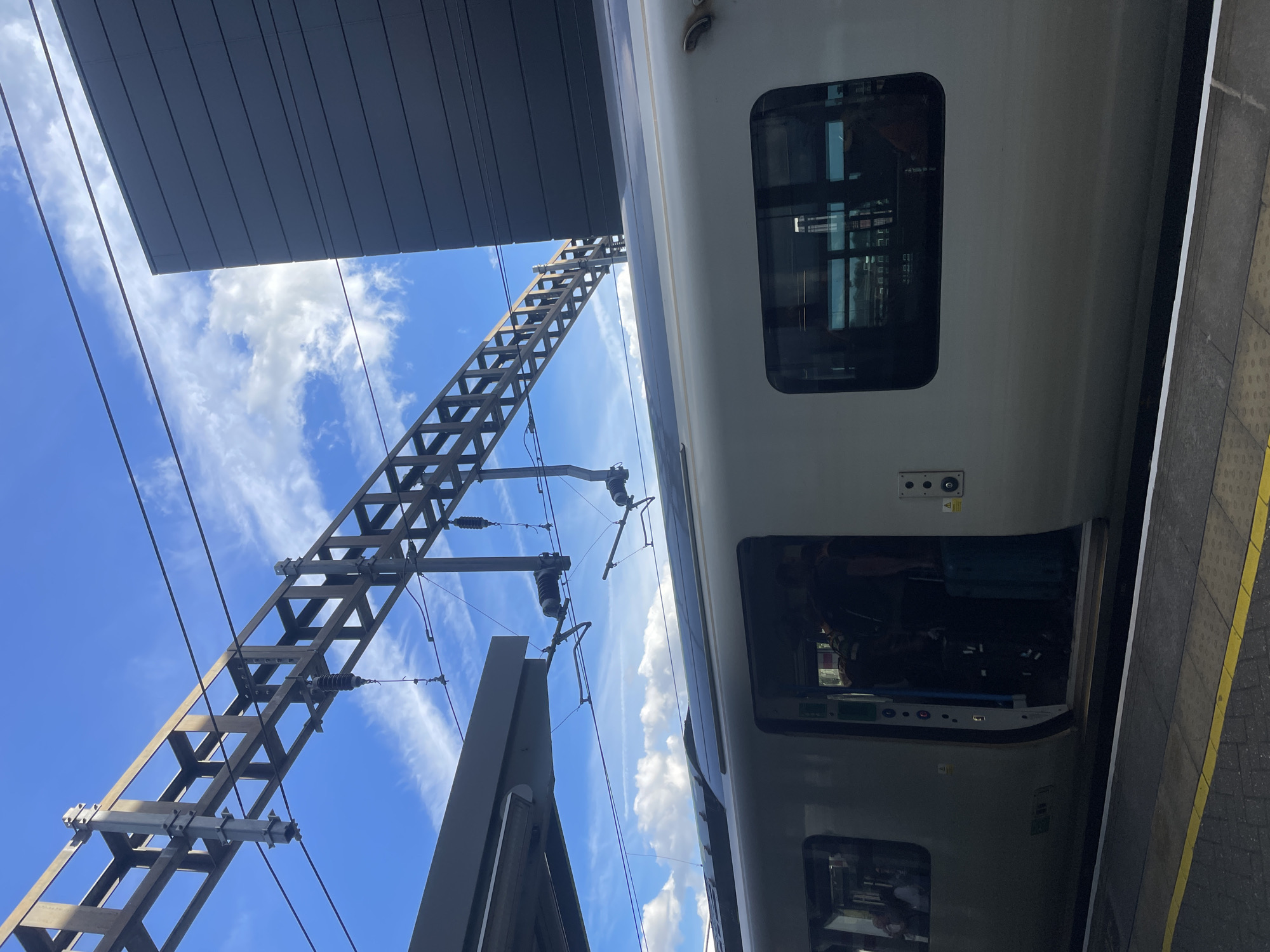 An exterior, low-angle, full shot captures a modern train at the Luton train station platform, under a bright blue sky with scattered white clouds. The train, a sleek, silver passenger carriage, has its doors open, revealing passengers boarding with luggage. The train's windows reflect the sky and the surrounding structures.
Above the train, overhead power lines and a complex metal support structure dominate the upper portion of the frame, indicative of an electrified railway. To the right, a contemporary building with dark, horizontally-paneled cladding rises, contrasting with the open sky and the industrial overhead infrastructure. The platform is visible in the foreground, with yellow and white tactile paving lines marking the edge. The scene appears to be captured on a clear day.