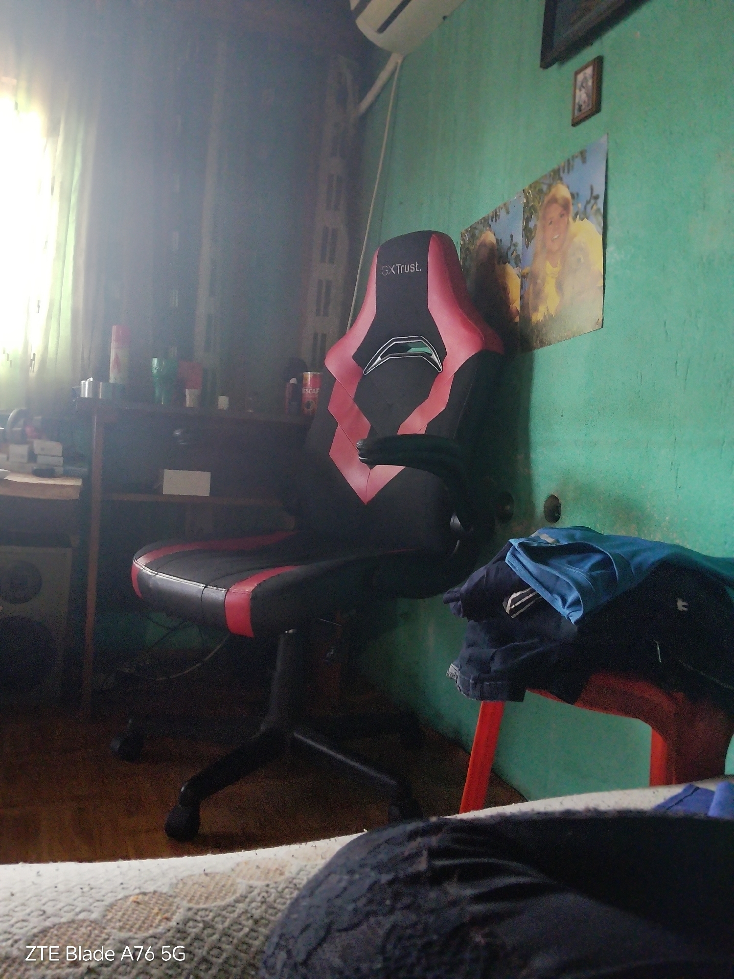 The image depicts an indoor residential room located in Stara Zagora, Bulgaria, illuminated by strong natural light entering from a window on the left, which is covered by light-colored curtains. The room features a prominent black and red "GXTrust" gaming chair positioned centrally. To the left of the chair, a dark wooden desk holds various items including a red canister, a green cup, and other containers. A "NESCAFÉ" can is visible on the right side of the desk. Below the desk, a shelf contains a white rectangular object, and a speaker unit is visible on the floor beneath the left side of the desk. The wall is painted a textured, light green/teal color. Adorning the wall are a framed picture in the upper right and a larger poster featuring a smiling blonde woman in a yellow top. An air conditioning unit is visible in the upper left corner of the wall. The floor is covered with wooden planks. In the foreground, a patterned rug or blanket partially obscures the lower part of the scene, and a person's legs, clad in dark clothing, are partially visible. A red plastic stool in the lower right corner supports a stack of folded garments, including a blue shirt and darker clothing items. No direct actions or interactions are depicted; the scene is static.
