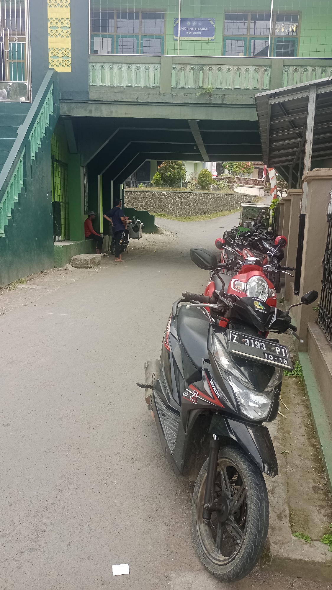 The image depicts a street scene in Karyamandala, Indonesia, featuring several motorcycles parked along the side of a road. In the foreground, a black and red Honda scooter is prominently displayed, with its license plate reading "Z 3193 PI 10-18". Behind it, a row of other motorcycles, predominantly red and black, are also parked. The background reveals a building with green walls and windows, identified by a sign as "SMK IBNU HAMBAL". A pedestrian overpass or walkway with green railings is visible above the street, connecting to the building. Two individuals are seen in the background, one sitting and the other standing near the building entrance. The environment suggests a daytime setting with overcast skies. The road is paved, and there is a stone wall bordering a small elevated area with some vegetation.