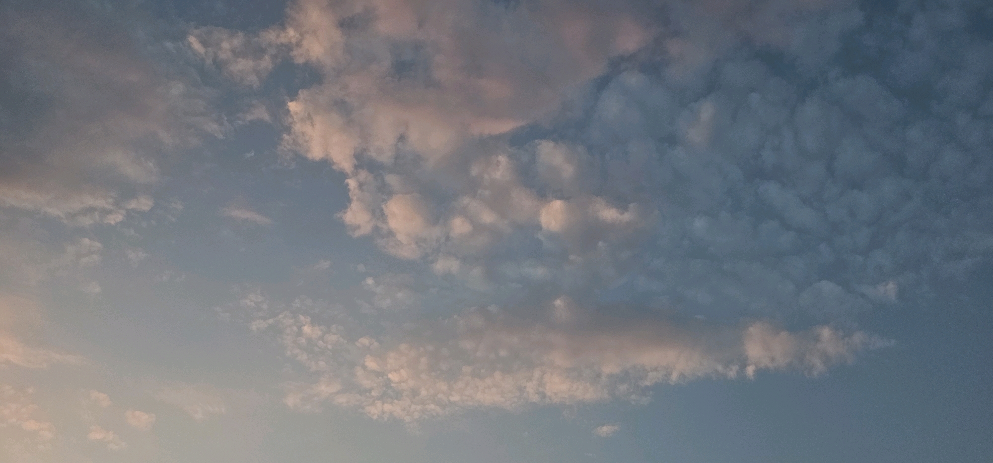 A wide-angle shot captures a soft, pastel sky filled with scattered clouds. The clouds exhibit a range of hues from pale pink and peach to a muted grey-blue. The lighting suggests either dawn or dusk, casting a gentle glow across the celestial scene. There are no discernible landmarks or man-made structures, and therefore no information about its location in Longqiao, China can be inferred. The image is solely focused on the atmospheric conditions.