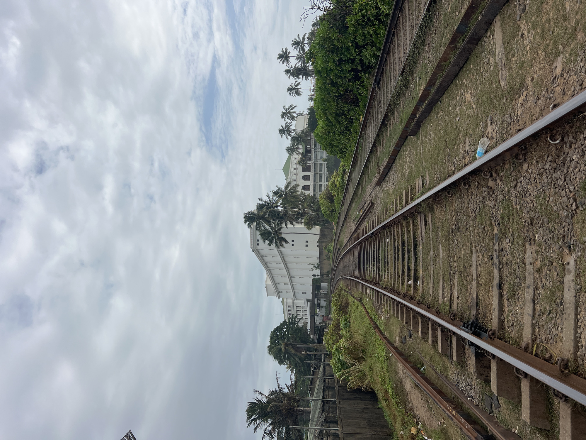 The image shows a set of parallel train tracks curving through a landscape. In the background, a large, white, multi-story building with a modern architectural style is visible, surrounded by lush green vegetation and palm trees. The sky is overcast with scattered clouds. The area around the tracks appears somewhat unkempt, with some debris visible on the left side. This scene is located in Dehiwala, Sri Lanka. There are no people or specific actions depicted.