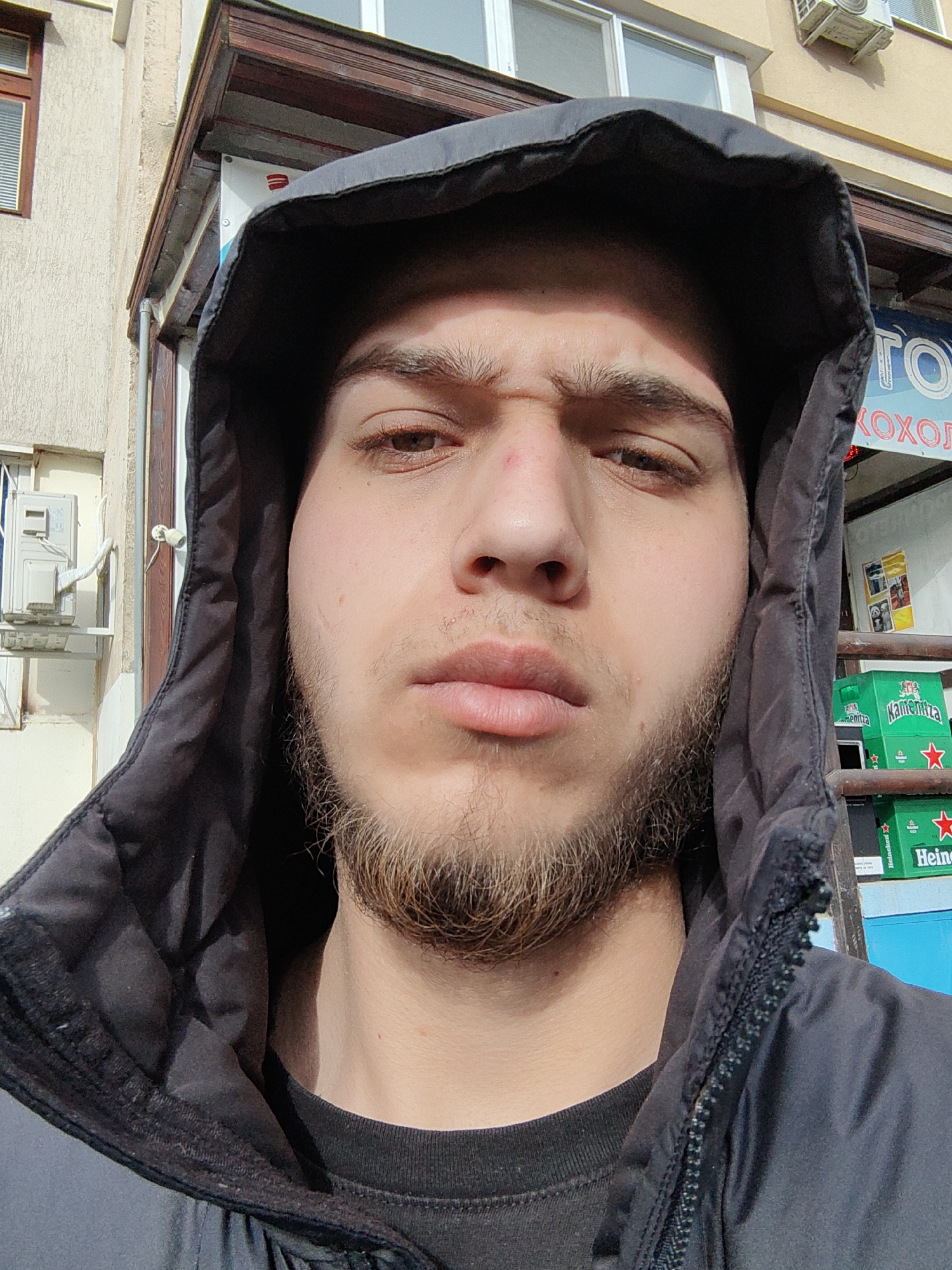 A close-up portrait of a young man with a light complexion, brown hair, and a beard. He is wearing a black hooded jacket, with the hood pulled up, partially obscuring the background.
The background includes a light-colored building with windows, an air conditioning unit, and a sign written in Cyrillic. Stacks of beer are visible in the background.
Based on the Cyrillic signage, the location is likely in Sofia, Bulgaria.