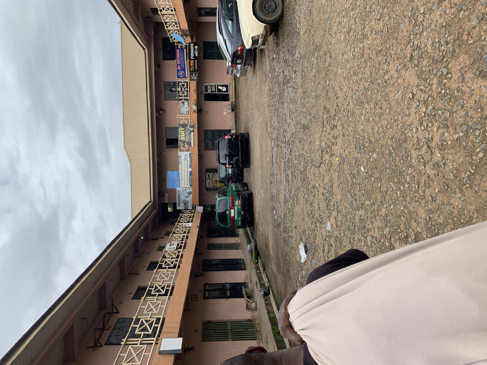 The image shows a courtyard with a two-story building surrounding it. The building appears to be a commercial complex with multiple shops and offices, each with a numbered entrance. There are several cars parked in the courtyard, including a green van with "AMBASSADOR 003" written on the back, a black SUV, and other passenger cars. Some of the shops have signs with business names such as "CELEBRITY," "JD RECORDS," and "WARR FASHIONS." The ground of the courtyard is covered with gravel and dirt, with some scattered debris. A person is visible in the foreground, facing away from the camera and holding a light-colored cloth. The sky is overcast. The scene is likely located in Jos, Nigeria, given the presence of a Nigerian license plate on one of the vehicles and the architectural style of the building.