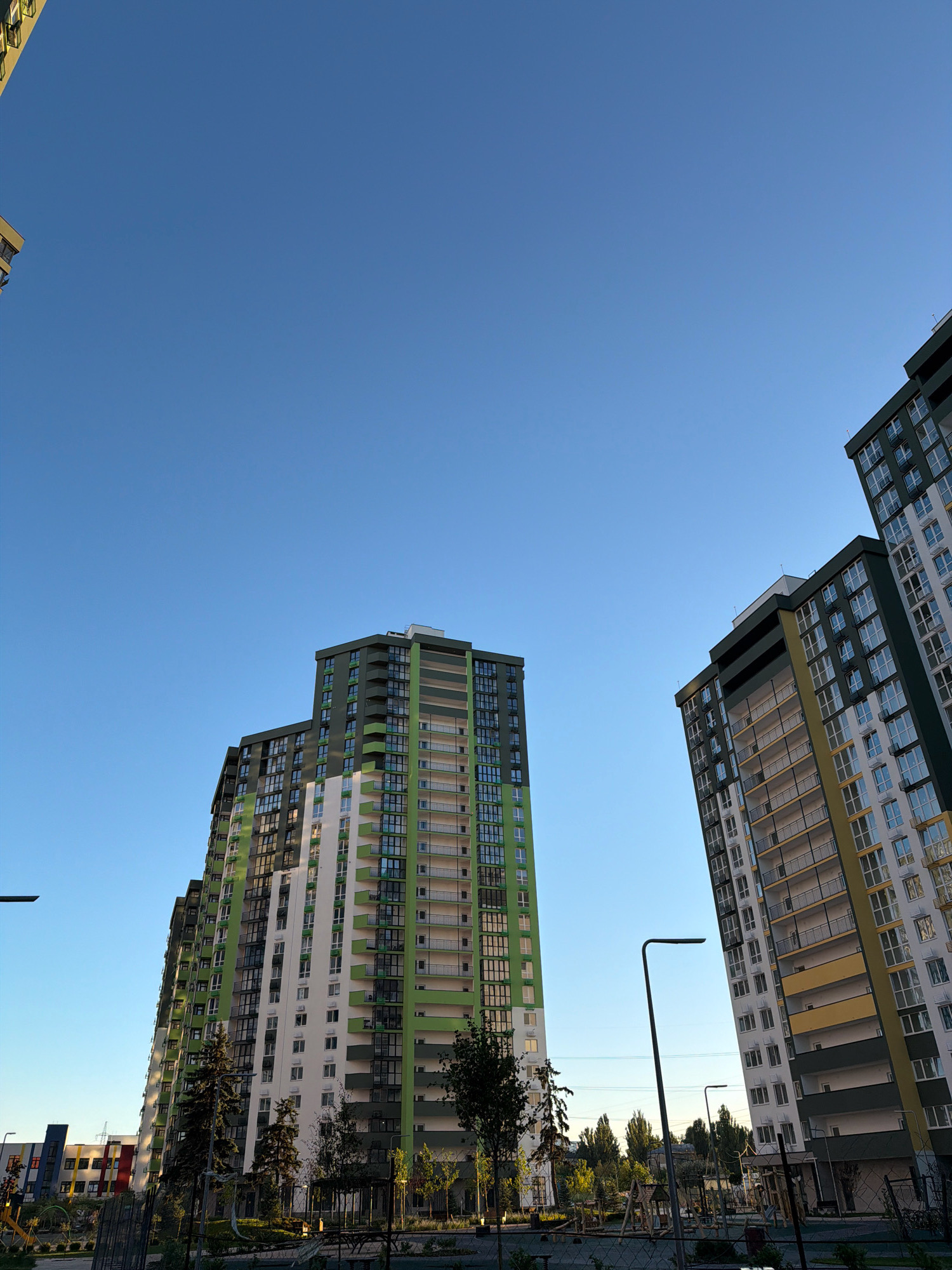 The image shows a clear blue sky over a residential complex in Kyiv, Ukraine. Several tall, modern apartment buildings with balconies and numerous windows dominate the foreground. The buildings feature a mix of white, green, and yellow facades. In the background, more buildings and trees are visible, suggesting a suburban or urban setting. A streetlamp stands in the middle ground. The scene appears to be taken during daylight, with shadows indicating the sun is relatively low in the sky, possibly late afternoon. There are no people or discernible actions depicted in the image. The overall impression is of a contemporary urban landscape under a vast sky.
