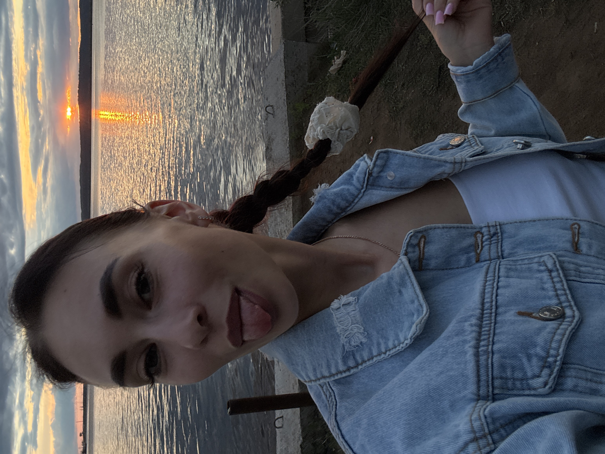 A young woman is posing for a selfie outdoors near a body of water, likely a lake or large river, at sunset. She is wearing a light blue denim jacket over a white top. Her dark hair is styled in a braid with a white, possibly makeshift, hair tie at the end. She is sticking out her tongue in a playful manner. The background shows a body of water reflecting the golden light of the setting sun. The location appears to be the city of Chaikovsky, Russia, evidenced by the sunset over a wide, flat body of water, similar to descriptions of the Kama River and associated reservoirs in the area. The photograph captures a casual, personal moment.