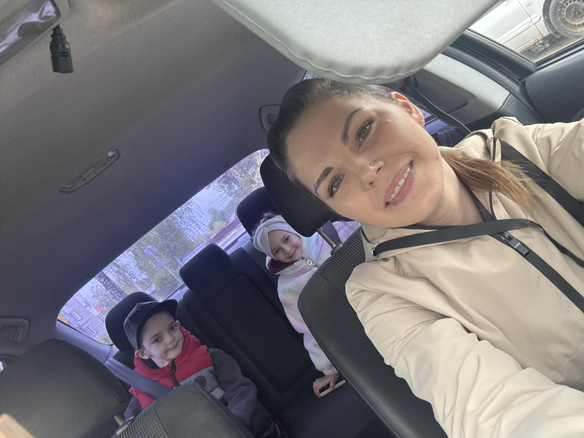 The image is taken from inside a car. In the front passenger seat is a woman with brown hair and braces, wearing a light-colored jacket and a black shoulder strap. In the back seats are two young children. One child, wearing a red jacket and a black cap, is in a car seat and buckled up. The other child, wearing a white hat and a pastel-colored jacket, is seated next to them. Outside the car window, there are buildings and trees, possibly indicating an urban or suburban environment. This image was taken in the city of Раздолье, Russia.