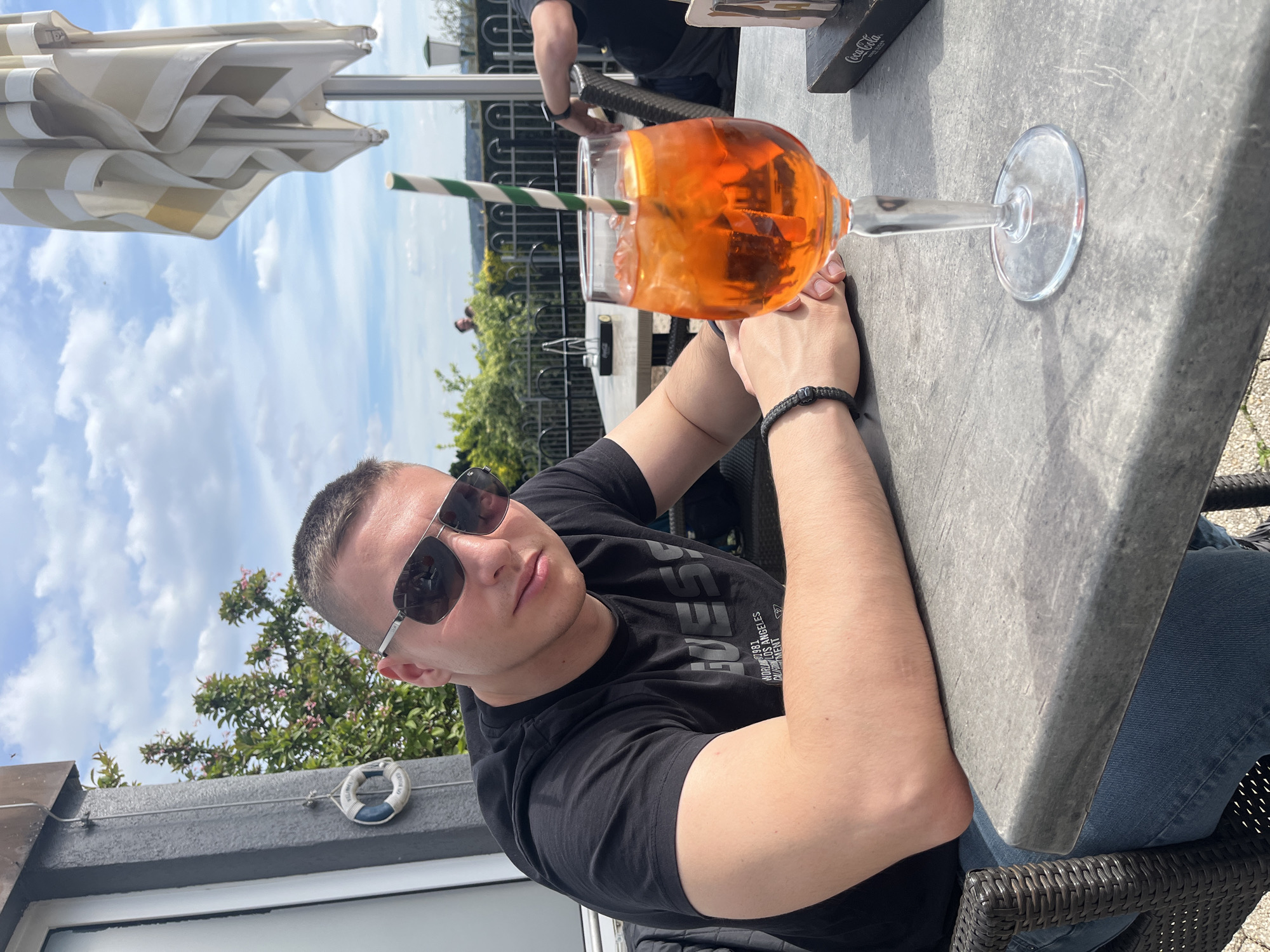 A man wearing sunglasses and a black t-shirt sits at a table in Saarburg, Germany. A glass of orange liquid with a green and white striped straw sits on the grey table next to his hands. A partially obscured umbrella is above, and trees and the sky can be seen in the background. A life preserver-like decoration is attached to a nearby structure. The man is looking at the camera. Another person, partially obscured, stands near the table.
