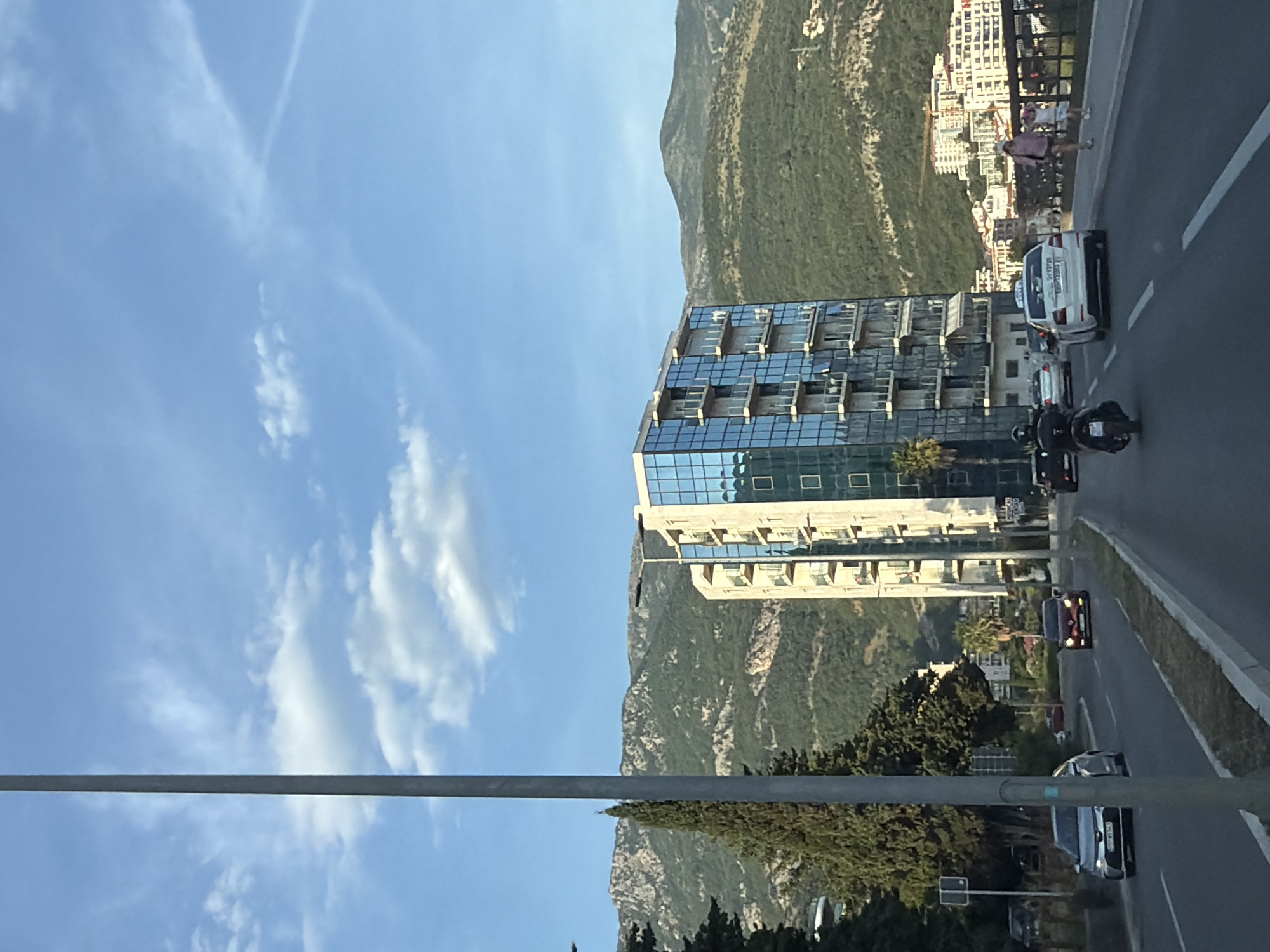 The image depicts a street scene in Boreti, Montenegro, on a sunny day with scattered clouds. A tall, modern building with a glass facade dominates the middle ground, reflecting the sky. In the background, a mountain covered in vegetation rises.
On the right side of the image, a road is visible with cars and motorcycles traveling along it. A few pedestrians are walking along the edge of the road.
A metal pole stretches across the lower part of the image, partially obstructing the view.