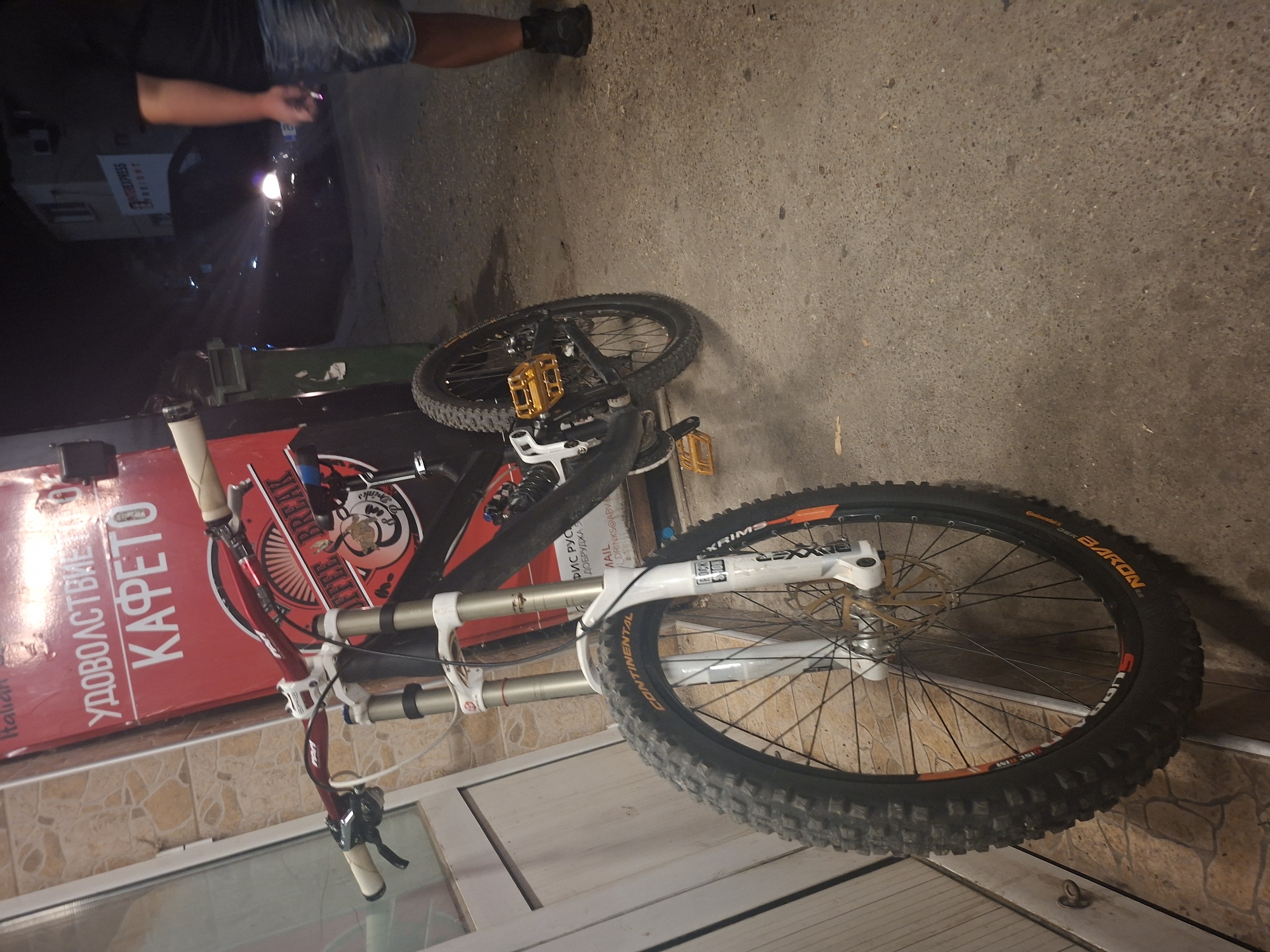 The scene is a nighttime outdoor view in Basarbovo, Bulgaria, featuring a downhill mountain bike prominently in the foreground.
The mountain bike, colored black and red, is equipped with a white RockShox BoXXer dual-crown suspension fork, gold flat pedals, and a red handlebar fitted with white grips and black brake levers. The front wheel displays a Continental Baron tire with aggressive tread and orange lettering, mounted on a black rim with a gold brake rotor. A similar Continental tire is visible on the rear wheel.
The bike leans against a building with reddish-brown, textured square wall tiles. A large red sign is affixed to this building, displaying white Cyrillic text "УДОВОЛСТВИЕ КАФЕТО" (Pleasure Coffee) and a "COFFEE BREAK" logo with a coffee cup graphic. Partial text "БИС РУС" and an email address "DRINKS@ABV.BG" are also visible on the lower portion of the sign. A white-framed door or window is partially visible on the far left.
The ground consists of a rough, grey paved surface, likely concrete or asphalt. In the middle ground, two garbage bins, one green and one darker, are positioned. Further in the background, a dark-colored vehicle with illuminated headlights is visible, partially obscured by darkness. Another building in the distance displays a sign with "INTEREXPRESS FREIGHT" printed on it.
On the far right of the frame, a male individual is partially visible from mid-thigh down. He is wearing a black t-shirt, light blue denim shorts, and black shoes. He is walking away from the camera and holding a small, light-colored object in his right hand. The overall lighting suggests ambient artificial illumination characteristic of a nighttime urban or semi-urban environment.
