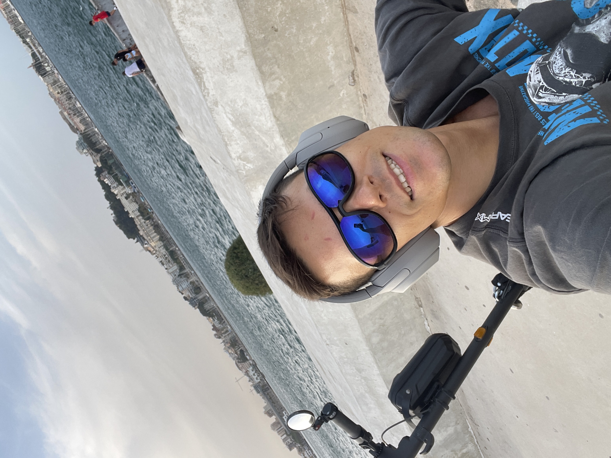 A man wearing headphones and sunglasses takes a selfie on a sunny day. He is seated on an electric scooter near a body of water, possibly the Mediterranean Sea, with a city skyline in the background. The city appears to be Cartagena, Spain, based on the architectural style and port activity visible. The man's reflection is visible in his sunglasses. He is smiling and wearing a dark gray t-shirt. In the background, several people are visible near the water's edge. The environment is bright and appears to be a promenade or walkway.