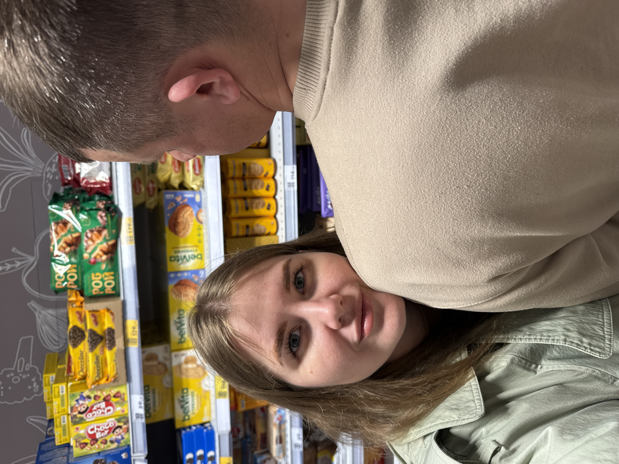 A woman with fair skin, light brown hair, and blue eyes poses for a photograph in a grocery store. She wears a light green jacket. Behind her, shelves are stocked with various snack products, including "belVita" breakfast biscuits, "Choco Boy" cookies, and "Рос-Пром" biscuits. In front of her is the back of a man’s head and shoulder. The setting is identifiable as a grocery store, as evidenced by the products and shelving. The location is the city of Горный, Russia.