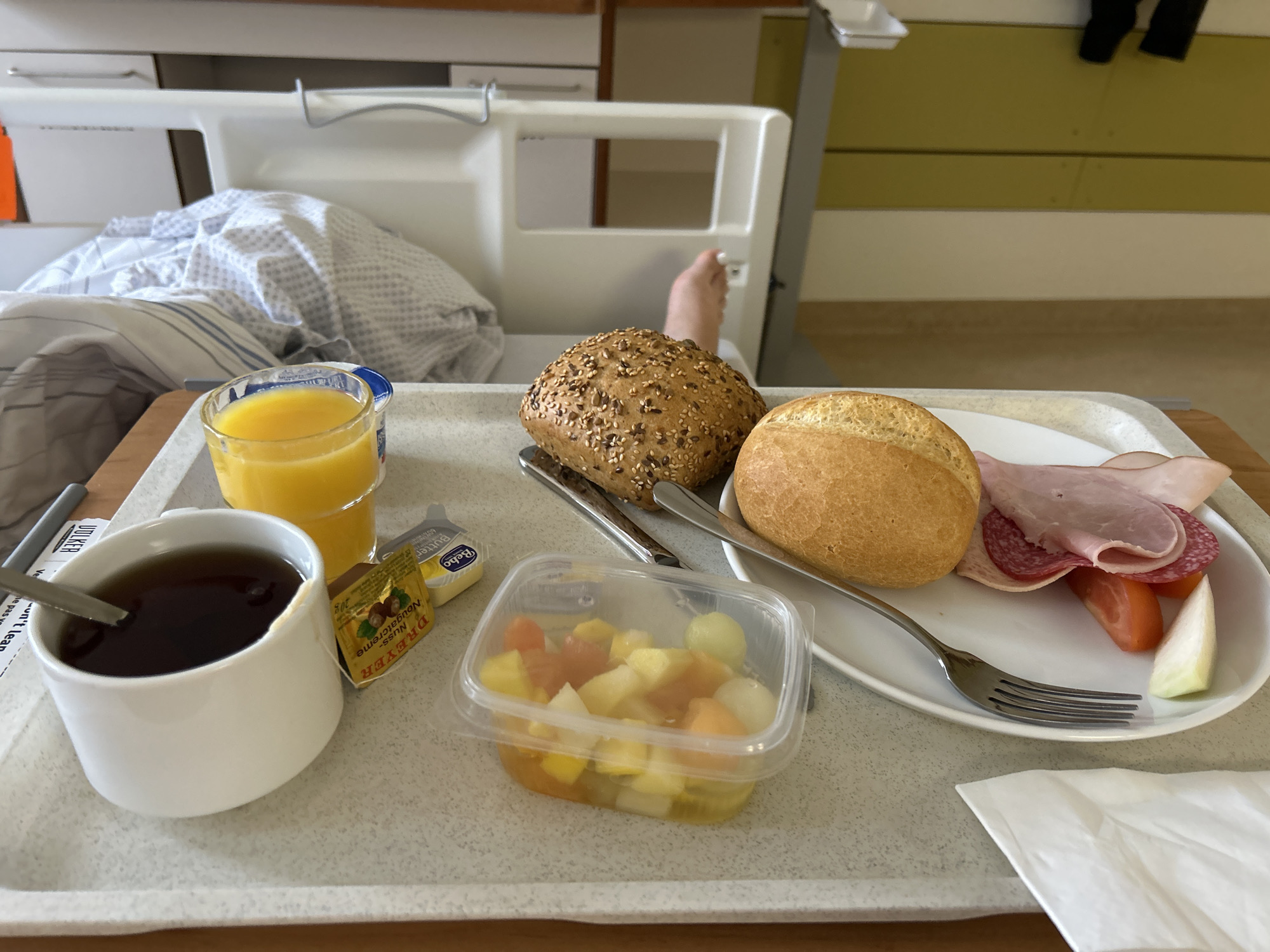 This is a photograph of a breakfast tray in a hospital room. The tray contains a cup of black tea, a glass of orange juice, a small tub of butter, a package of hazelnut spread, a container of diced fruit salad, a round bread roll with seeds, a plain bread roll, a slice of ham, a slice of salami, a slice of tomato, and a slice of onion. A fork rests on the plate with the meats and vegetables. In the background, a hospital bed with a patient covered by a patterned blanket is visible, along with a portion of the patient's foot. The environment appears to be a hospital room with a beige and green striped wall in the background and a bedside cabinet. The scene is lit by ambient light.
