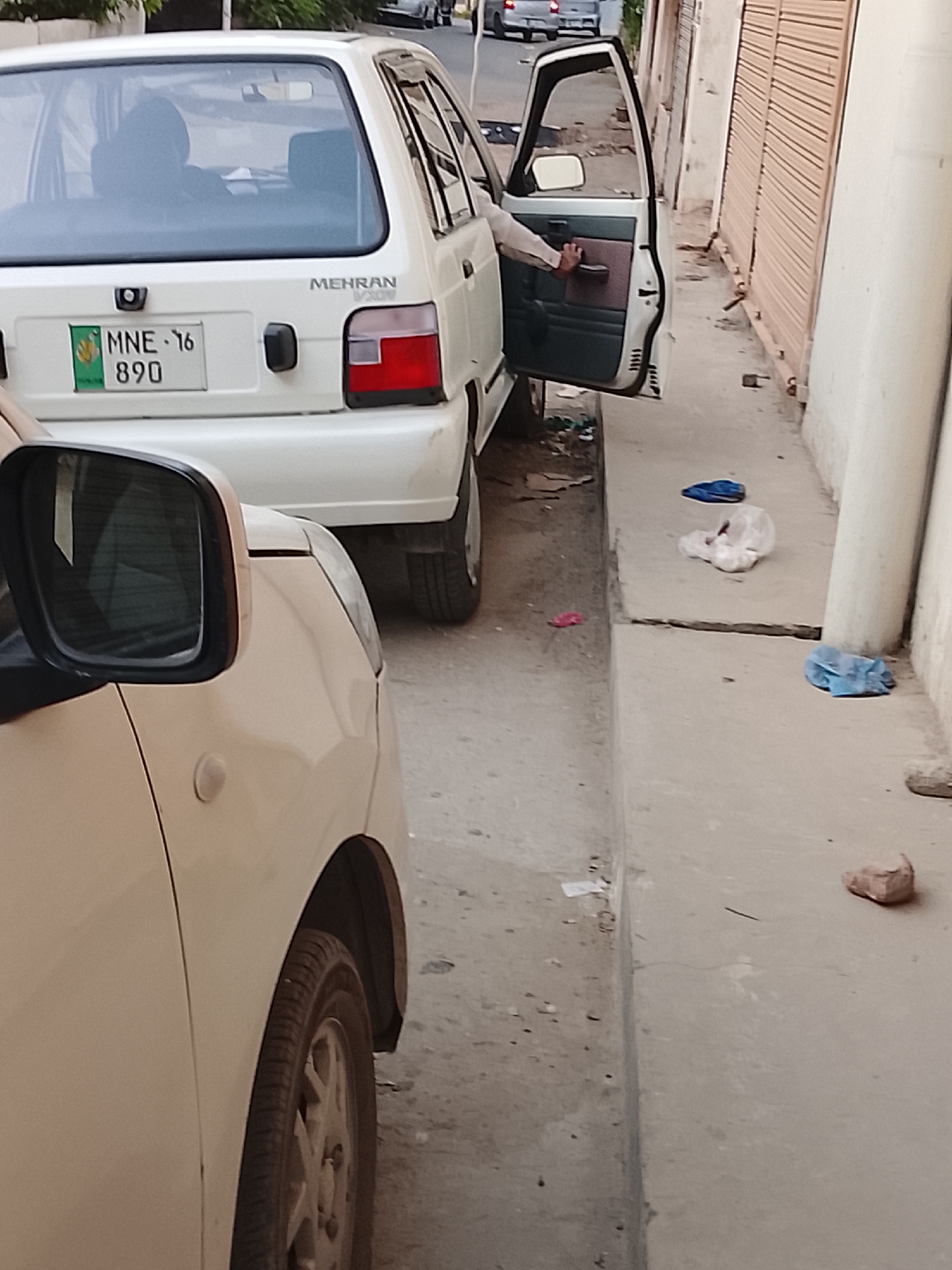 A white Suzuki Mehran car with the license plate "MNE-16 890" is parked partially obstructing a narrow street. A person's arm is visible, suggesting they are opening the driver-side door. In the foreground, a beige car partially blocks the view of the street, with its side mirror prominently displayed. The setting is an urban street with a narrow sidewalk cluttered with debris, including plastic bags and fabric scraps. Buildings with closed metal roll-up doors line the street. The image appears to be taken in Gulgasht Colony, Pakistan.